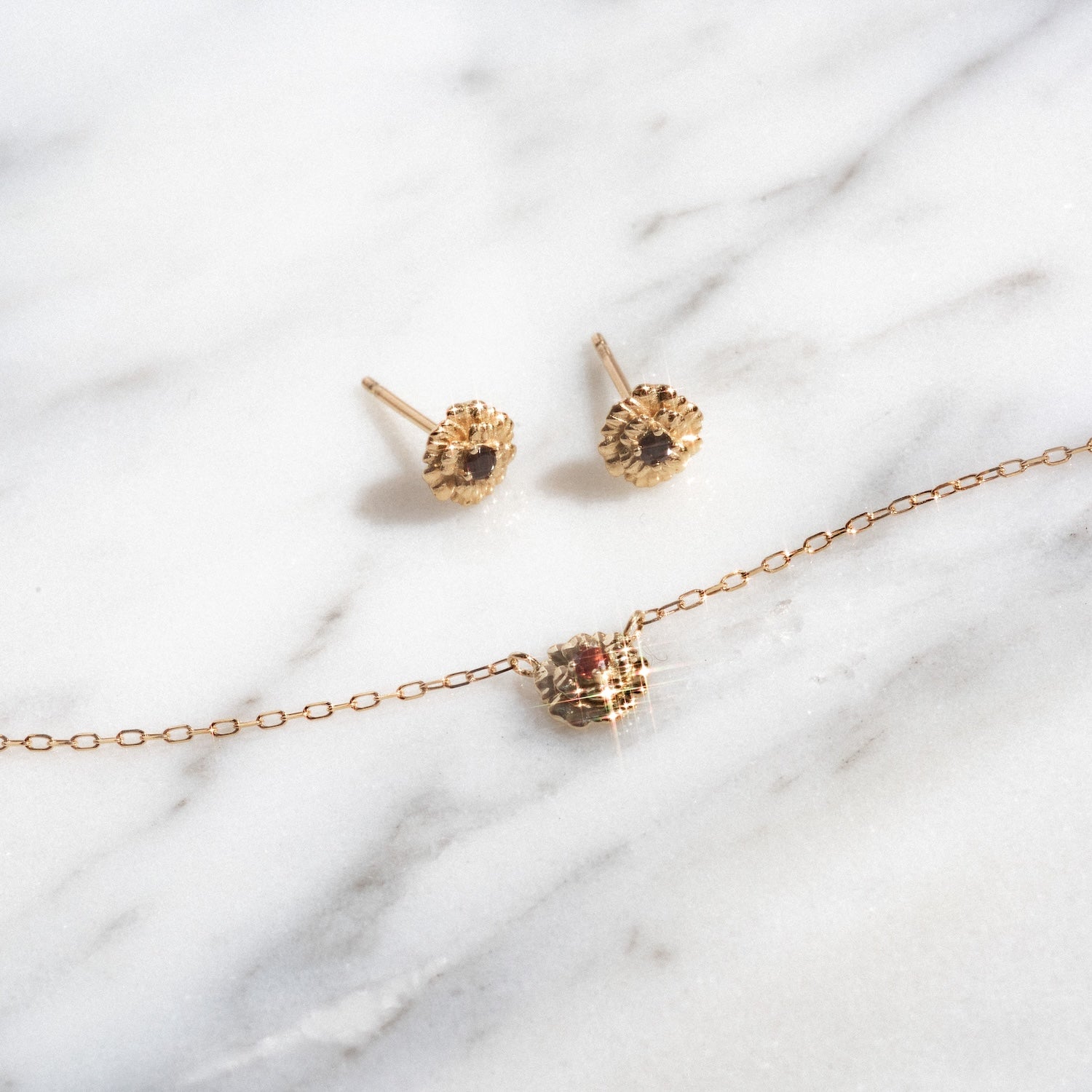 The Solid Gold Birth Flower Gift Set features gold stud earrings with dark central stones and a dainty gold chain necklace with a small red gemstone pendant, all displayed on a white marble surface.