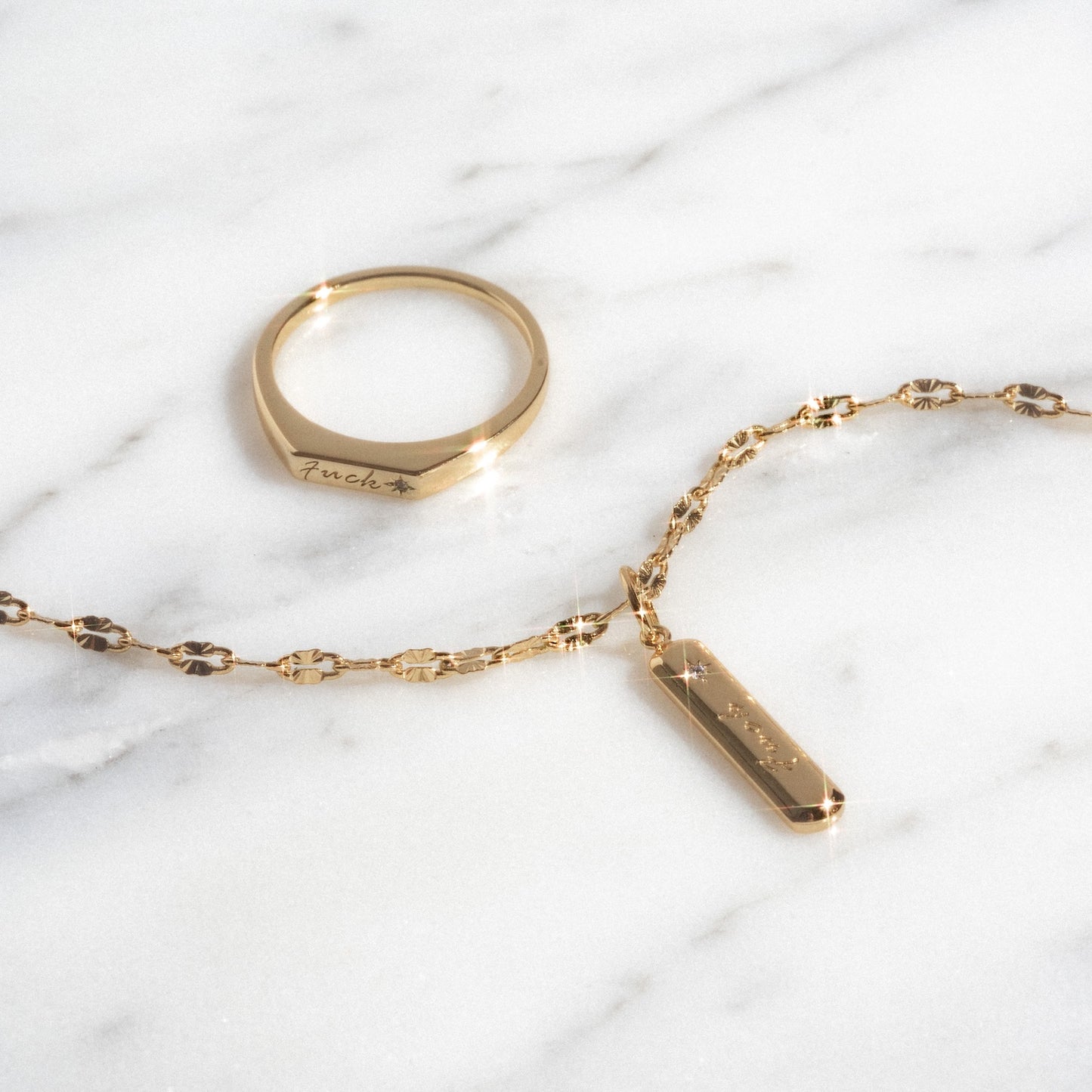 The Tiny Fuck Gift Set, featuring a gold ring and a 14k gold plated chain bracelet with a rectangular engraved pendant, is displayed on white marble. Both pieces catch the light to create subtle sparkles.