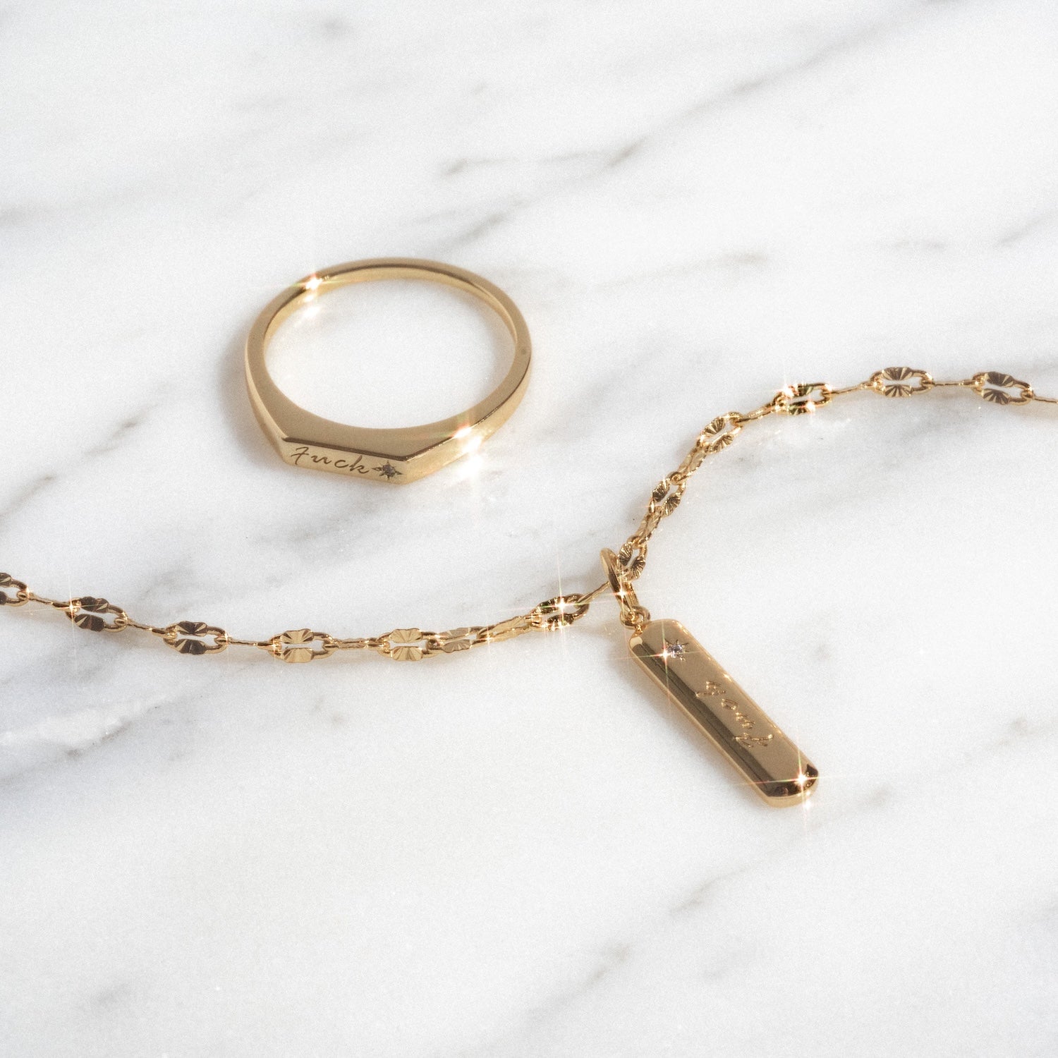 The Tiny Fuck Gift Set, featuring a gold ring and a 14k gold plated chain bracelet with a rectangular engraved pendant, is displayed on white marble. Both pieces catch the light to create subtle sparkles.