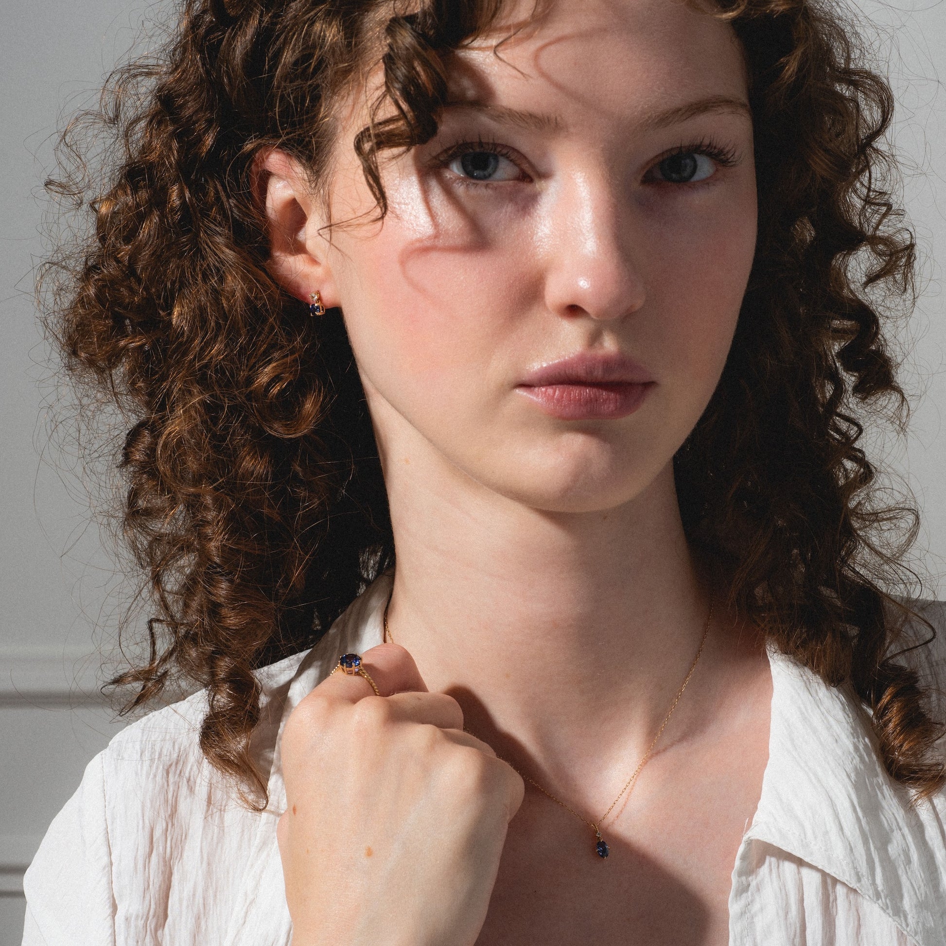 A young woman with curly brown hair wears a white shirt and the Solid Gold 2025 December Capsule Classic Tanzanite Pendant Necklace, gazing at the camera with a neutral expression against a light background.