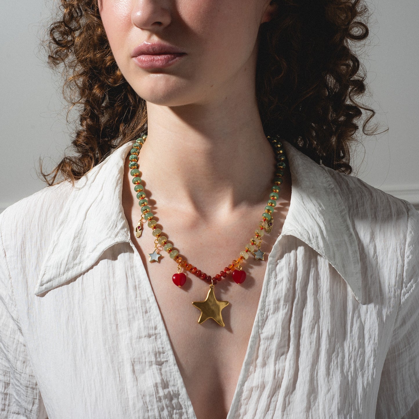 A woman with curly brown hair wears a white textured shirt and the True North Necklace, a colorful beaded piece with a large gold star and red heart charms, illuminated by soft natural light.