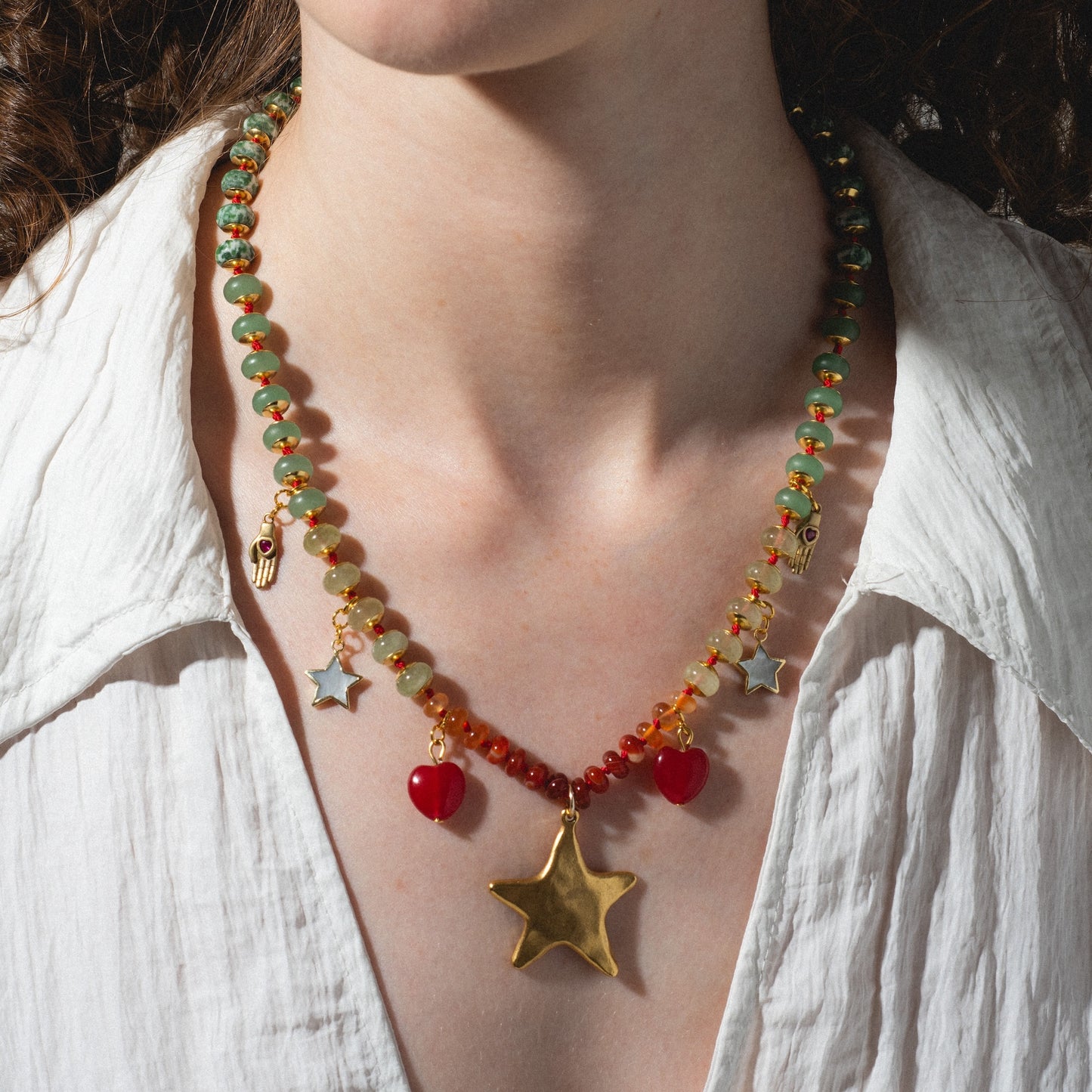 A person wearing a white shirt showcases the True North Necklace, featuring colorful green and red beads and gold charms, including a standout star pendant and charming heart accents.