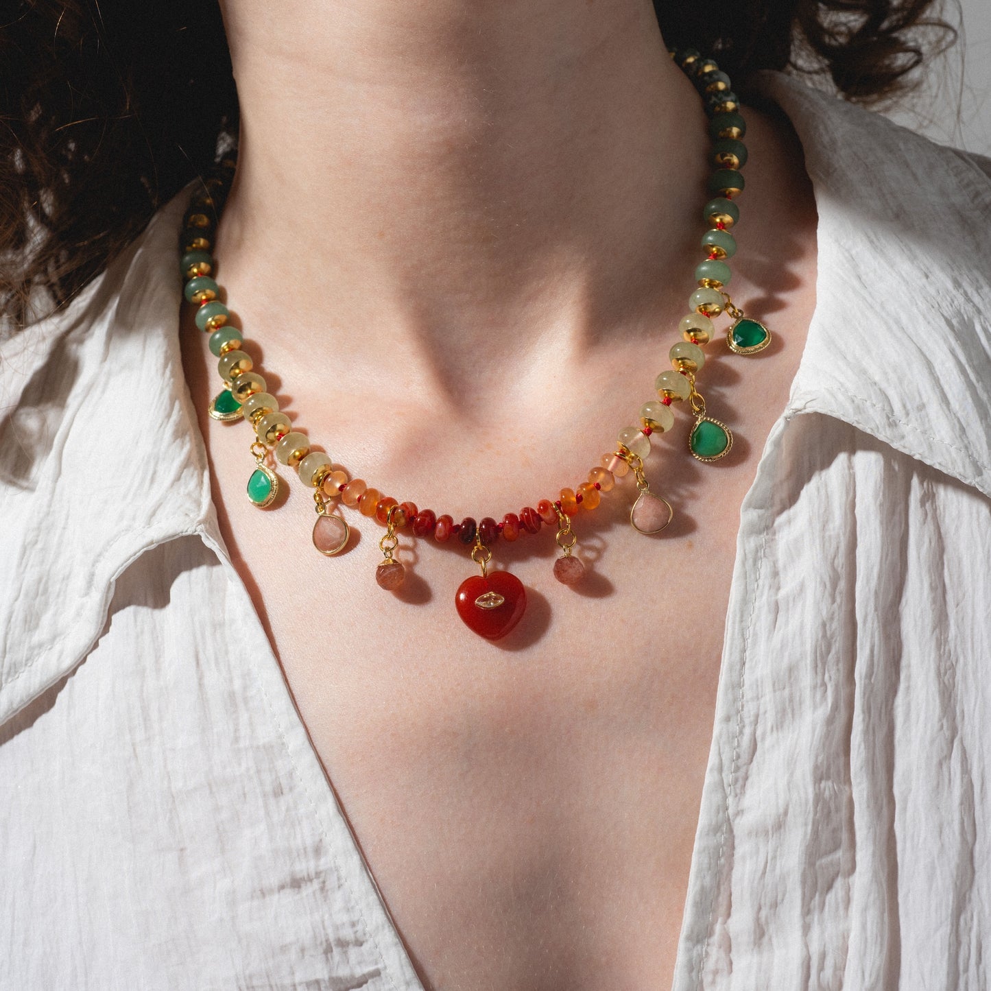 A person wearing a white shirt showcases the Golden Hour Necklace, featuring red, green, and yellow beads with dangling charms. The shot highlights their neck and chest.