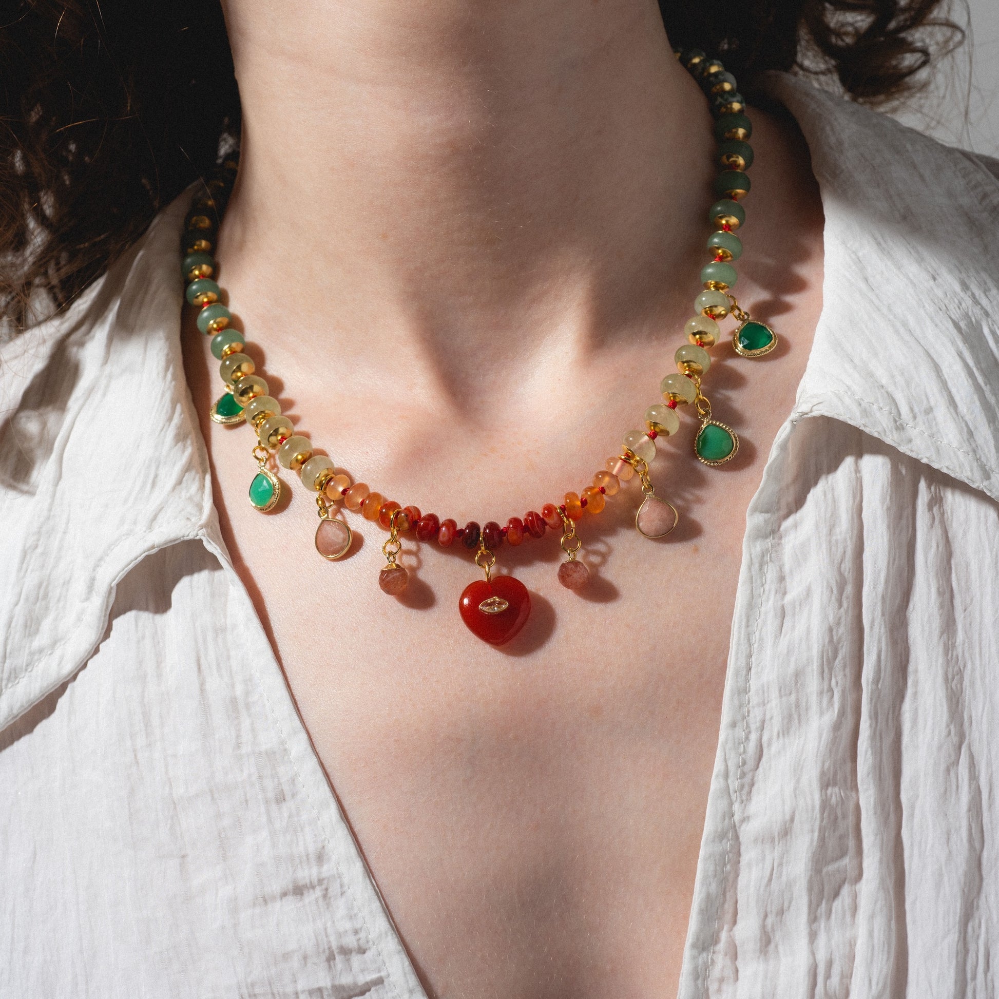 A person wearing a white shirt showcases the Golden Hour Necklace, featuring red, green, and yellow beads with dangling charms. The shot highlights their neck and chest.