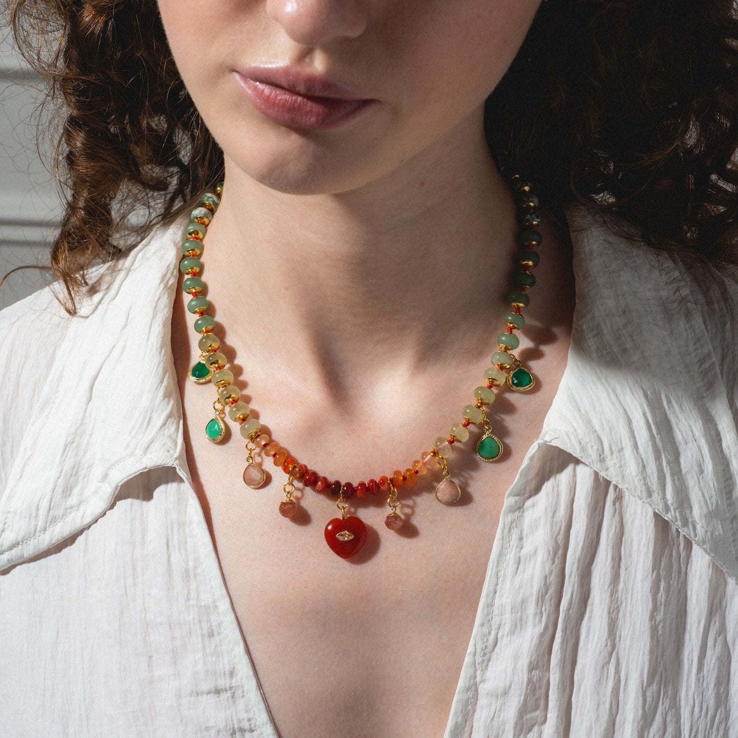 A woman with curly brown hair wears a white shirt and the Golden Hour Necklace, a colorful beaded accessory with green, red, and orange stones, shown from the shoulders up against a neutral background.