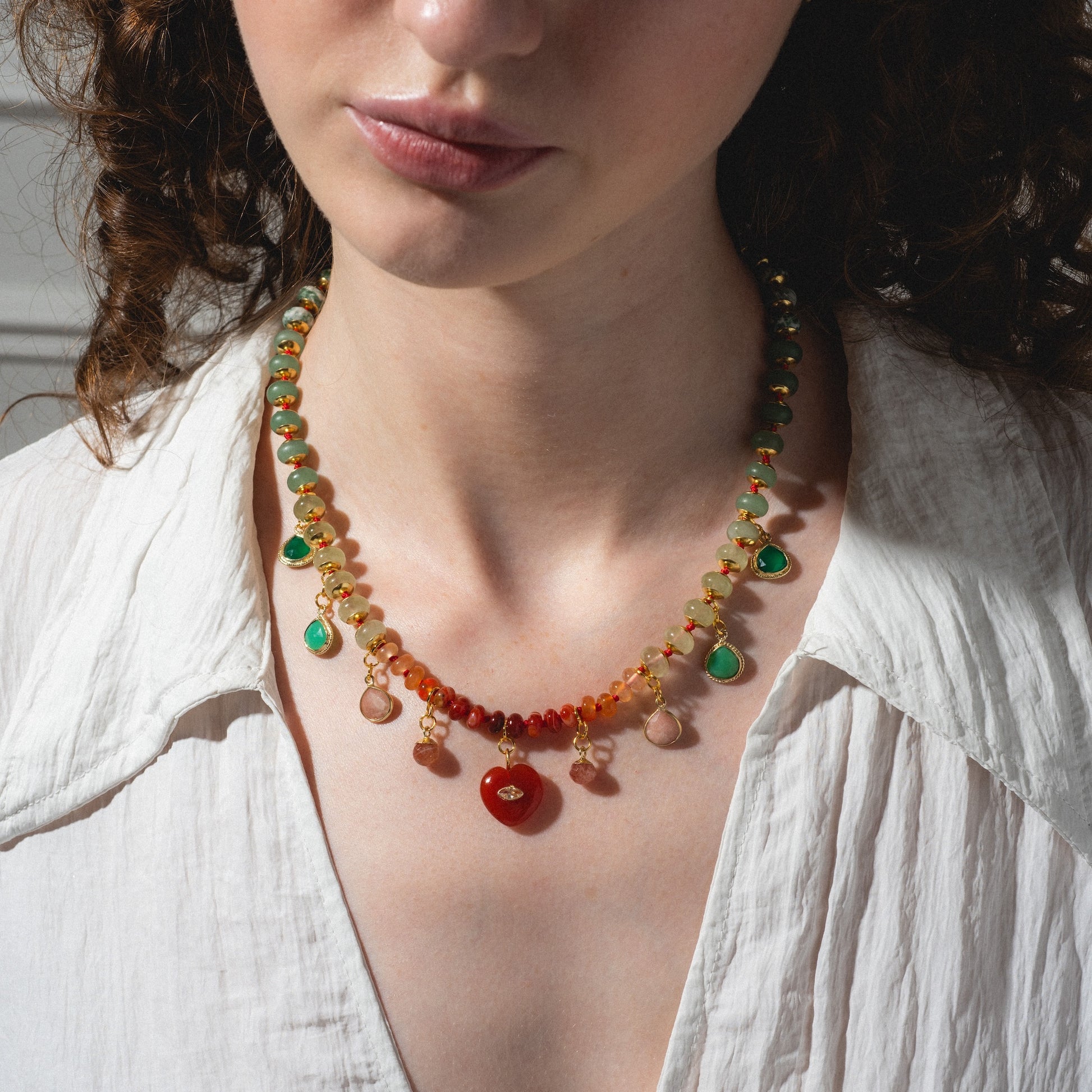 A woman with curly brown hair wears a white shirt and the Golden Hour Necklace, a colorful beaded accessory with green, red, and orange stones, shown from the shoulders up against a neutral background.