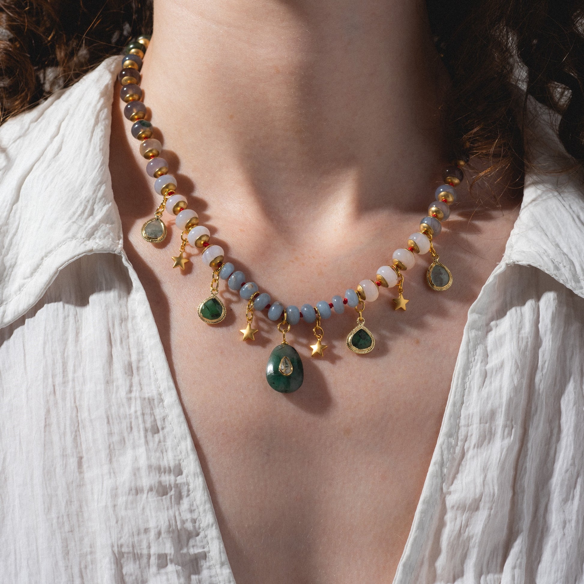 A person wearing the Winter Garden Necklace—pastel beads with green gemstones, gold stars, dangling gold-edged charms, and decorative pendants—paired with a white, open-collared shirt.