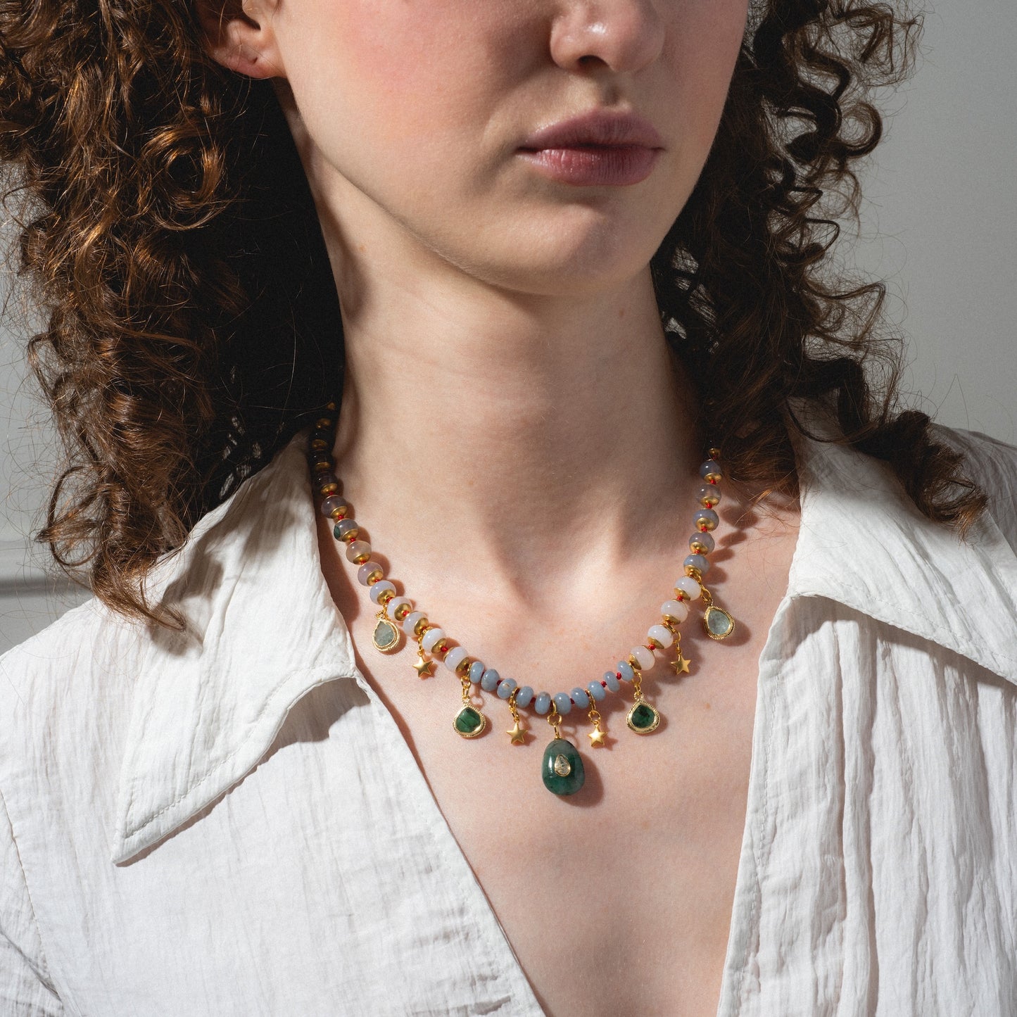 A woman with curly brown hair wears a white shirt and the Winter Garden Necklace, featuring green, gold, and blue beaded pendants. The image is cropped to show her neck and the lower part of her face.