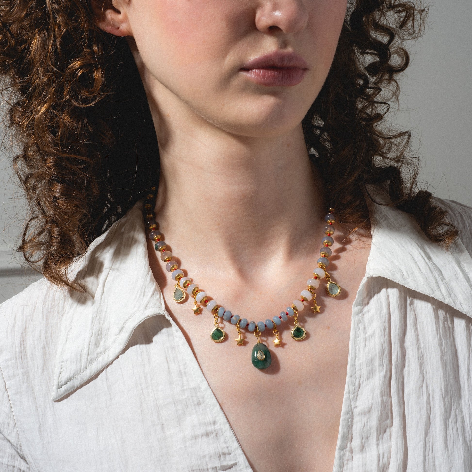A woman with curly brown hair wears a white shirt and the Winter Garden Necklace, featuring green, gold, and blue beaded pendants. The image is cropped to show her neck and the lower part of her face.