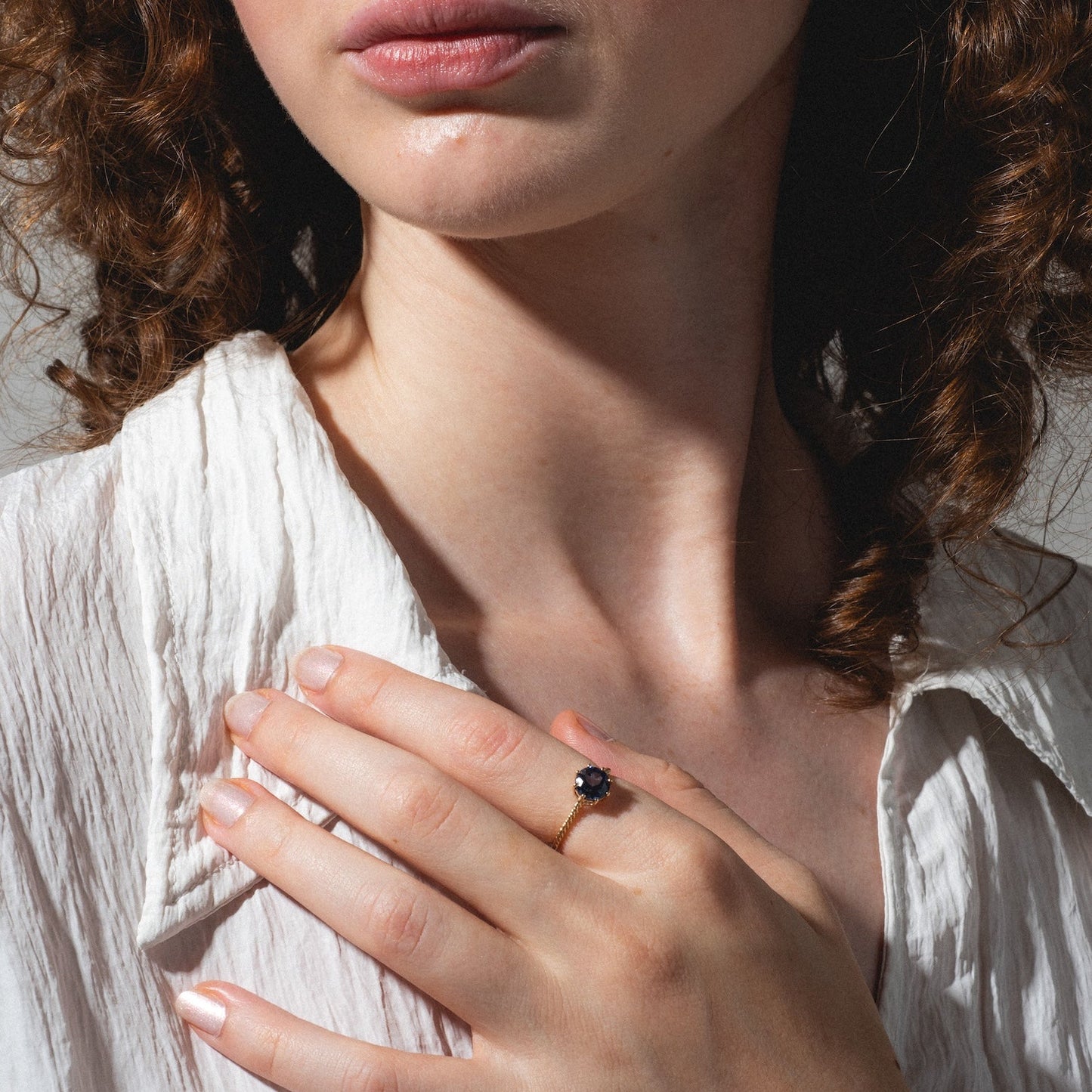 A woman with curly brown hair in a textured white blouse rests her hand on her chest, showcasing the Solid Gold 2025 December Capsule Classic Tanzanite Statement Ring. Light and shadow accentuate the ring, her face, and hand.