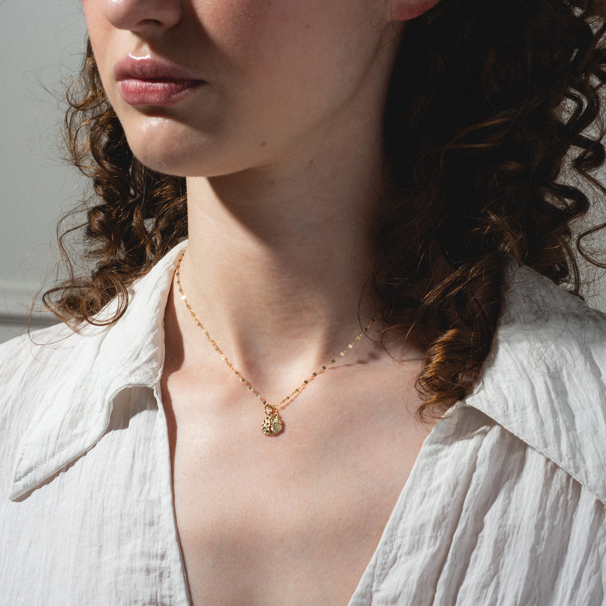 A woman with curly brown hair wears a white blouse and the Levain Bakery x Local Eclectic Solid Gold Share a Cookie Friendship Charm necklace. The image highlights her neck and the delicate gold pendant against a softly lit background.