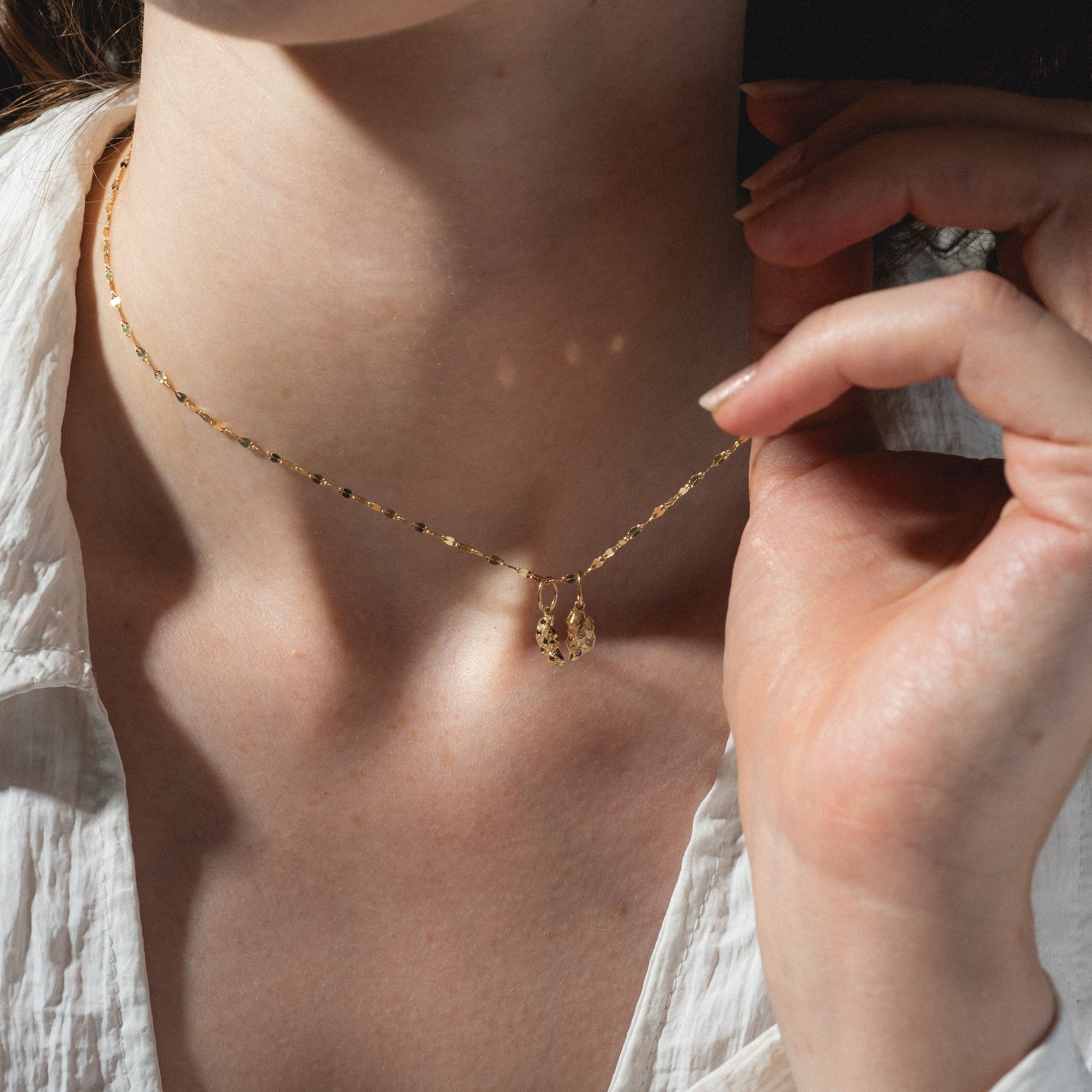 A person in a white shirt holds the Levain Bakery x Local Eclectic Solid Gold Share a Cookie Friendship Charm necklace, showcasing its delicate pendant against their neck in natural light.