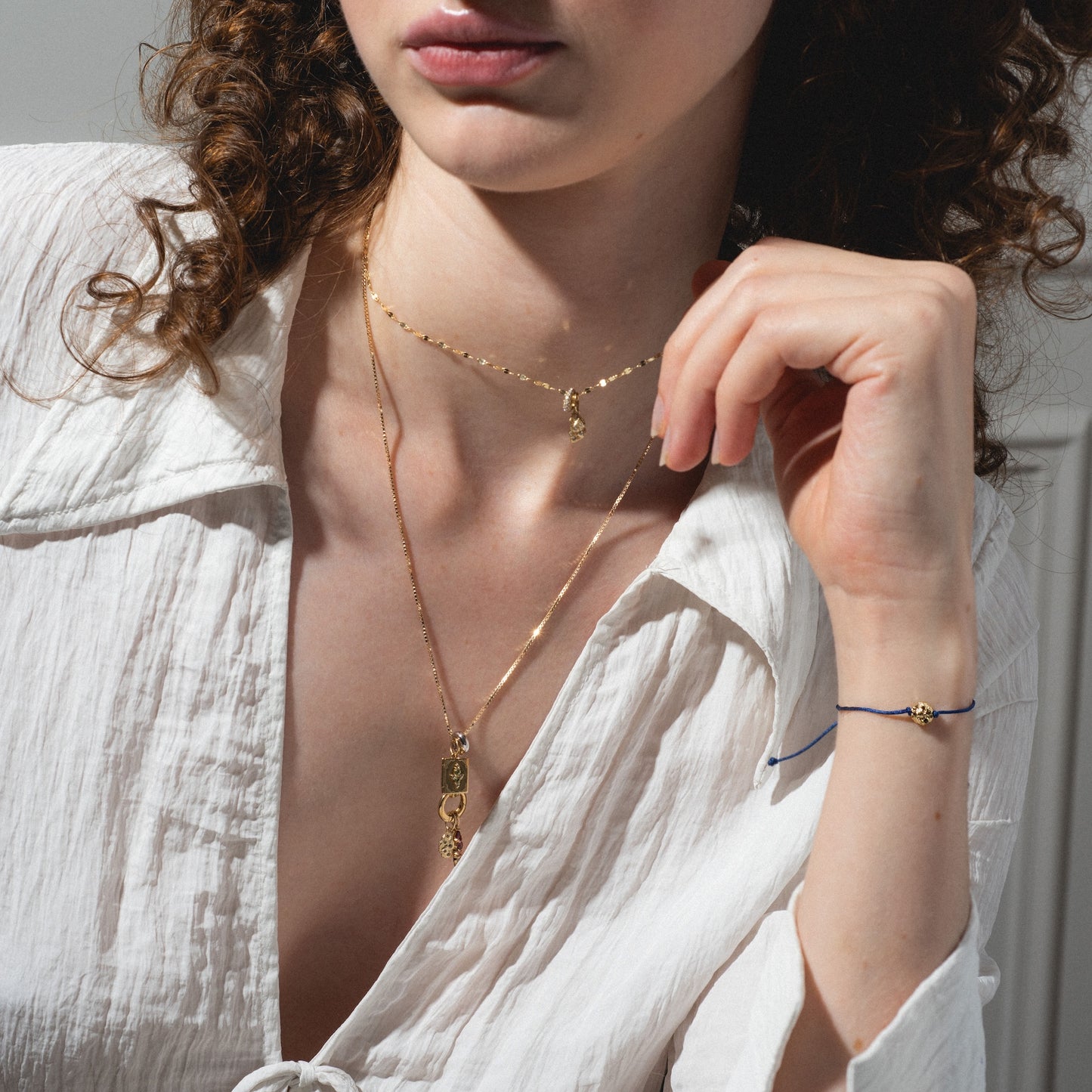 A woman in a white textured blouse wears layered gold necklaces and a delicate blue bracelet featuring the Levain Bakery x Local Eclectic Solid Gold Share a Cookie Friendship Charm. Her curly hair frames her face as she poses, highlighting the jewelry.