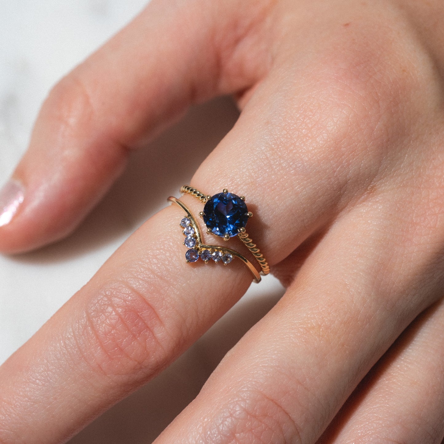 A hand wears two Solid Gold 2025 December Capsule Tanzanite rings: one with a large, round blue tanzanite gemstone and another featuring small purple stones in a chevron shape, included in the Full Set with Free Gift.