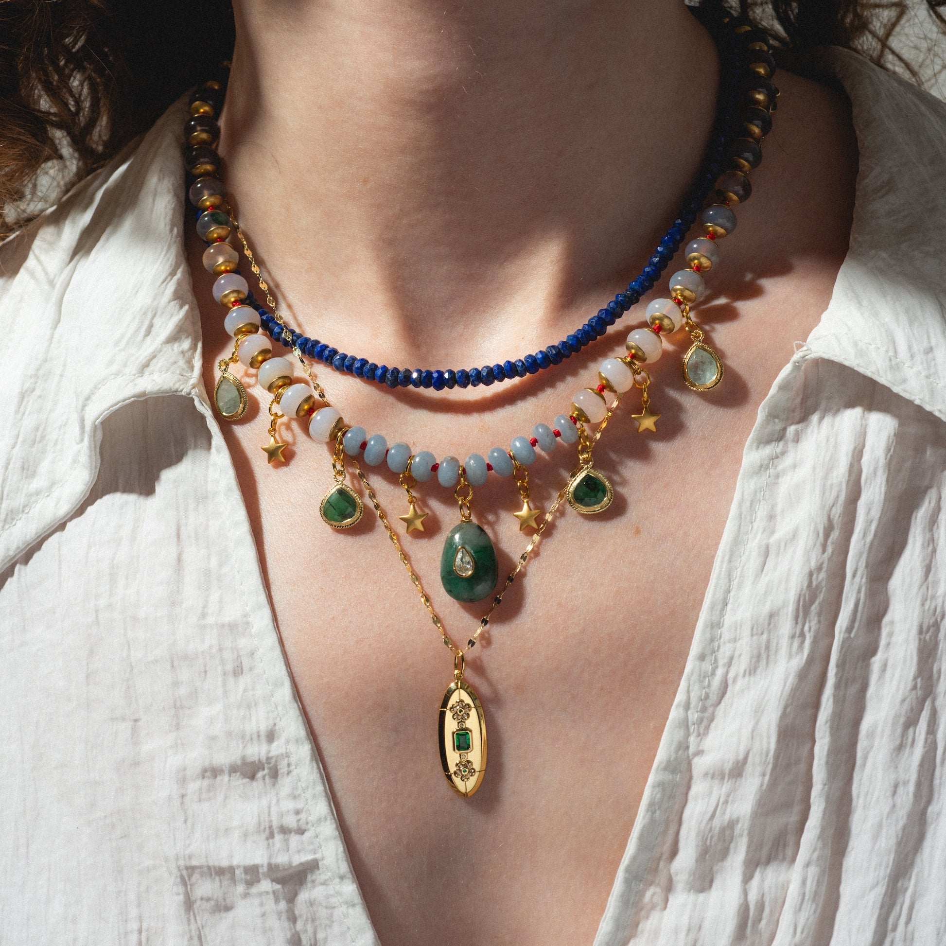 A person in a white shirt showcases the Winter Garden Necklace, which features three layers of blue and gold beads, green gemstones, gold star charms, and an ornate gold pendant with a green stone.