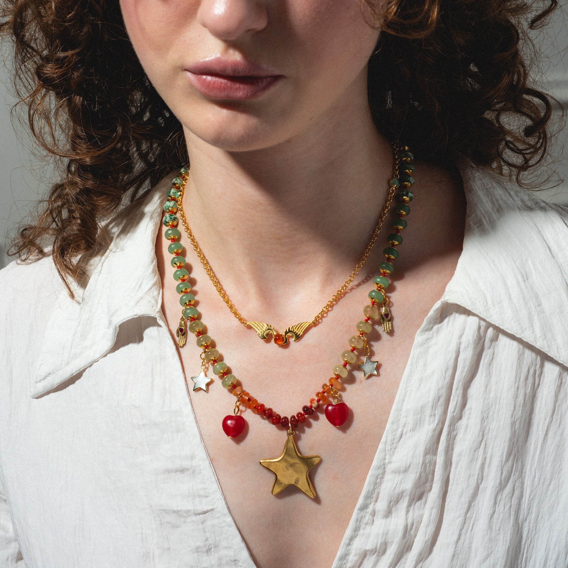 A woman in a white shirt wears the True North Necklace, featuring layered gold chains with a wavy design, colorful beads, a bold gold star pendant, smaller star charms, and red heart accents.