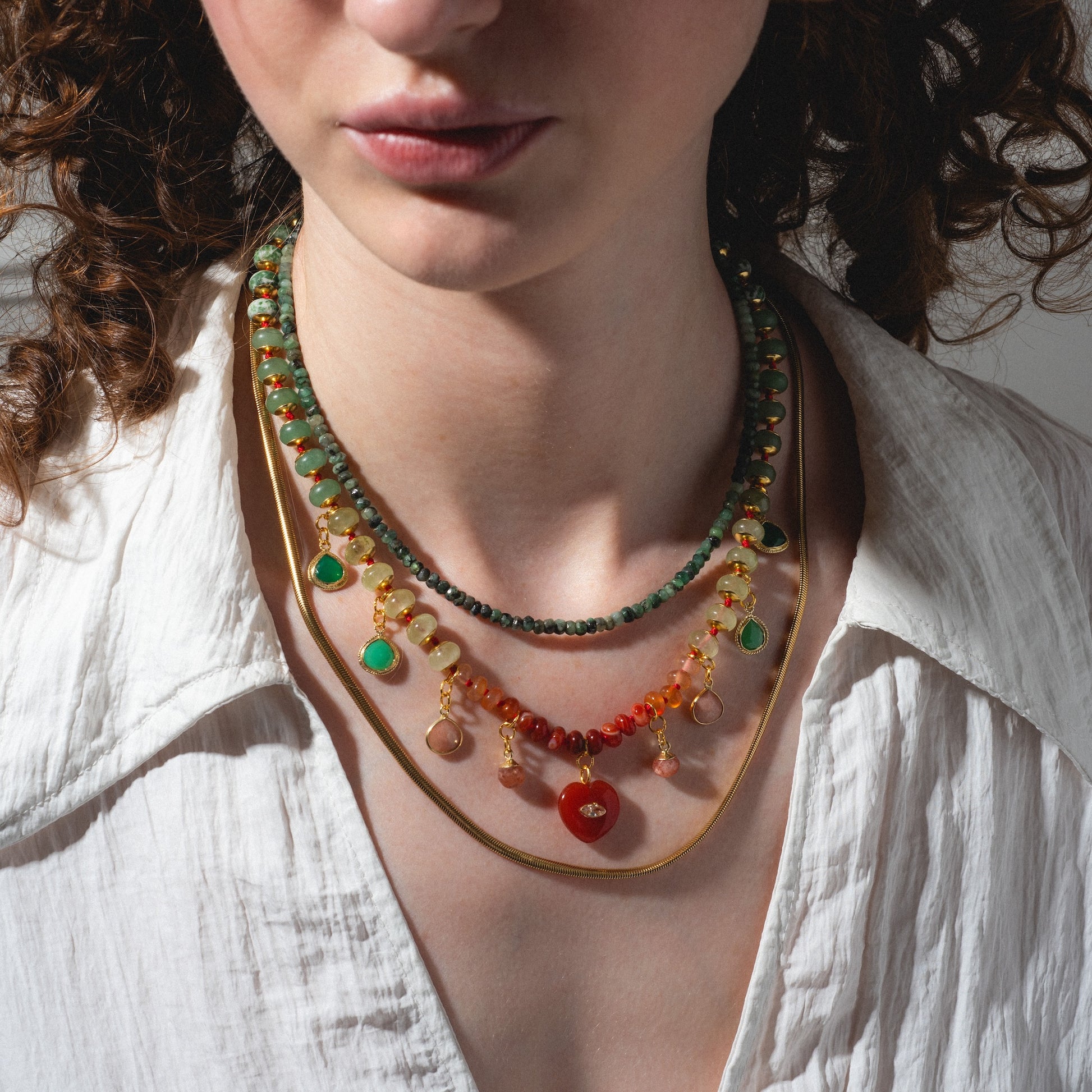 A person wears the Golden Hour Necklace—layered beads in green, red, and gold—over a white textured open-collar blouse. Only their lower face, neck, and upper chest are visible.