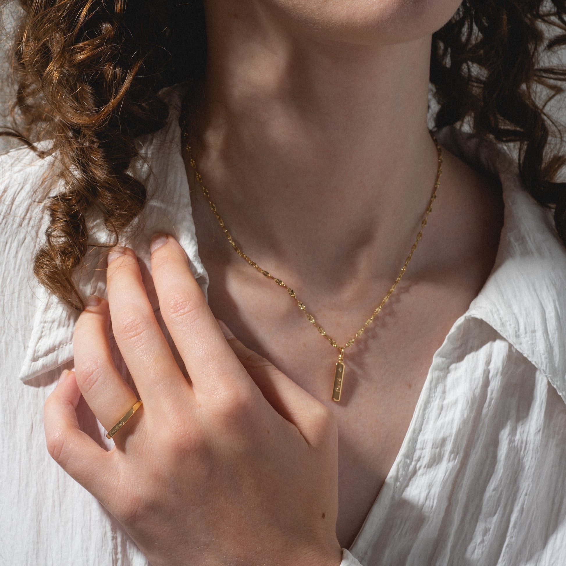 A person with curly brown hair wears a textured white shirt and the Tiny Fuck Gift Set—featuring a 14k gold plated rectangular pendant necklace and matching ring—lightly touching their collar for an effortlessly stylish look.