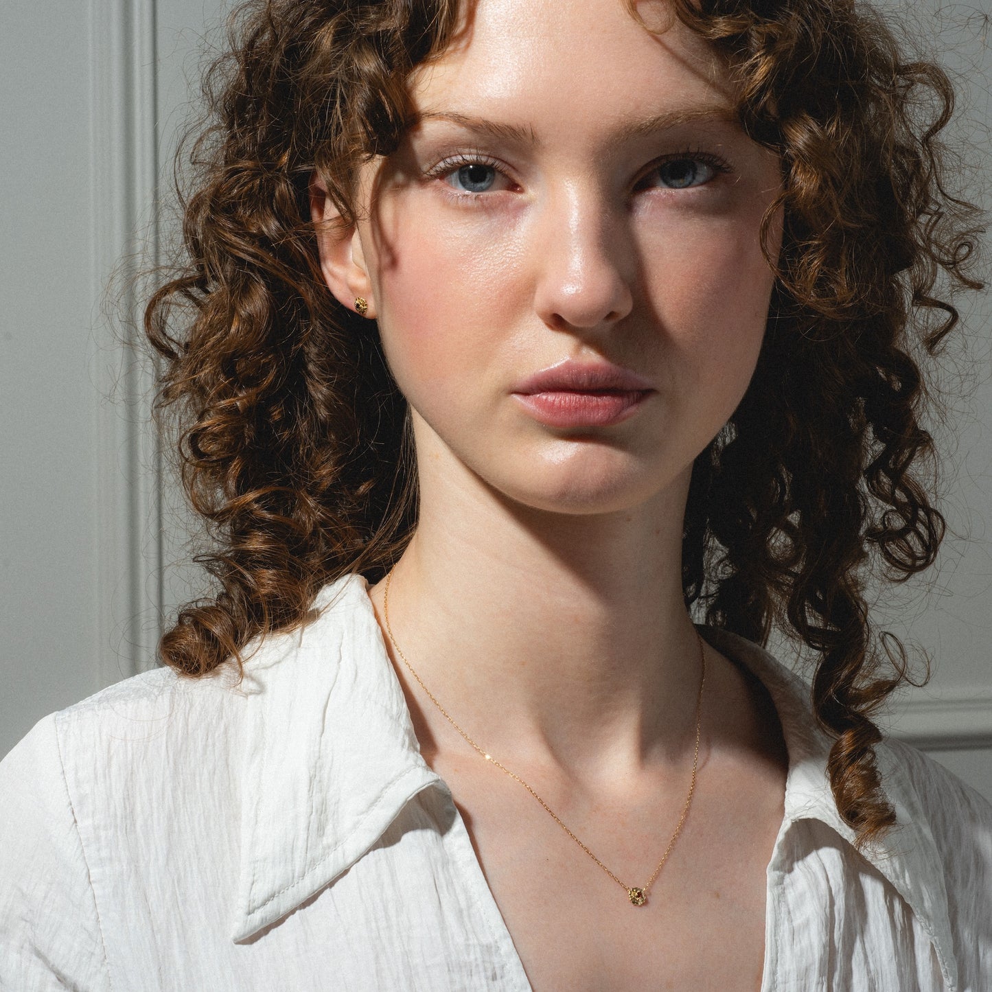 A young woman with curly brown hair and fair skin wears a white blouse and the Solid Gold Birth Flower Gift Set necklace, looking directly at the camera with a neutral expression against a softly lit background.