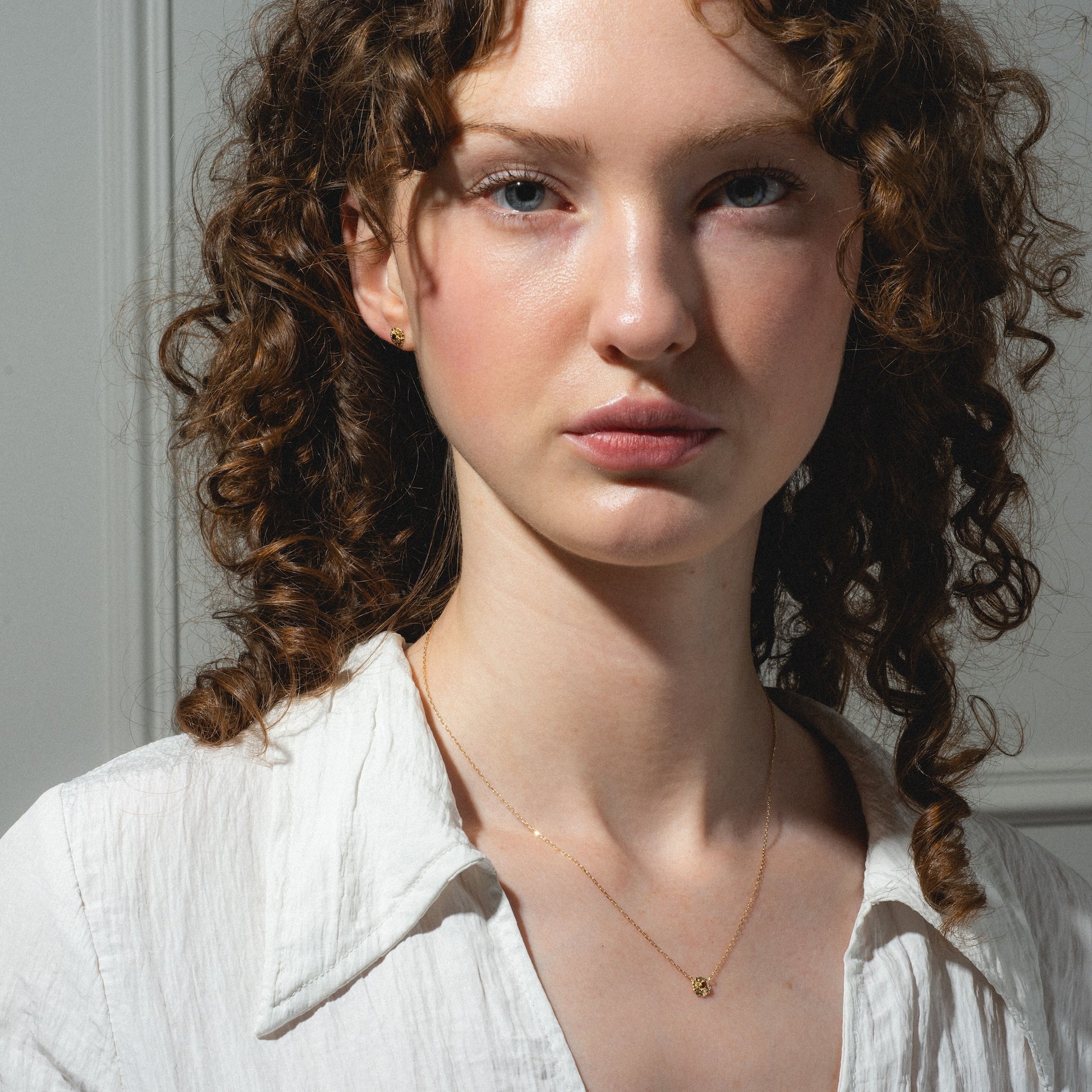 A young woman with curly brown hair and fair skin wears a white blouse and the Solid Gold Birth Flower Gift Set necklace, looking directly at the camera with a neutral expression against a softly lit background.