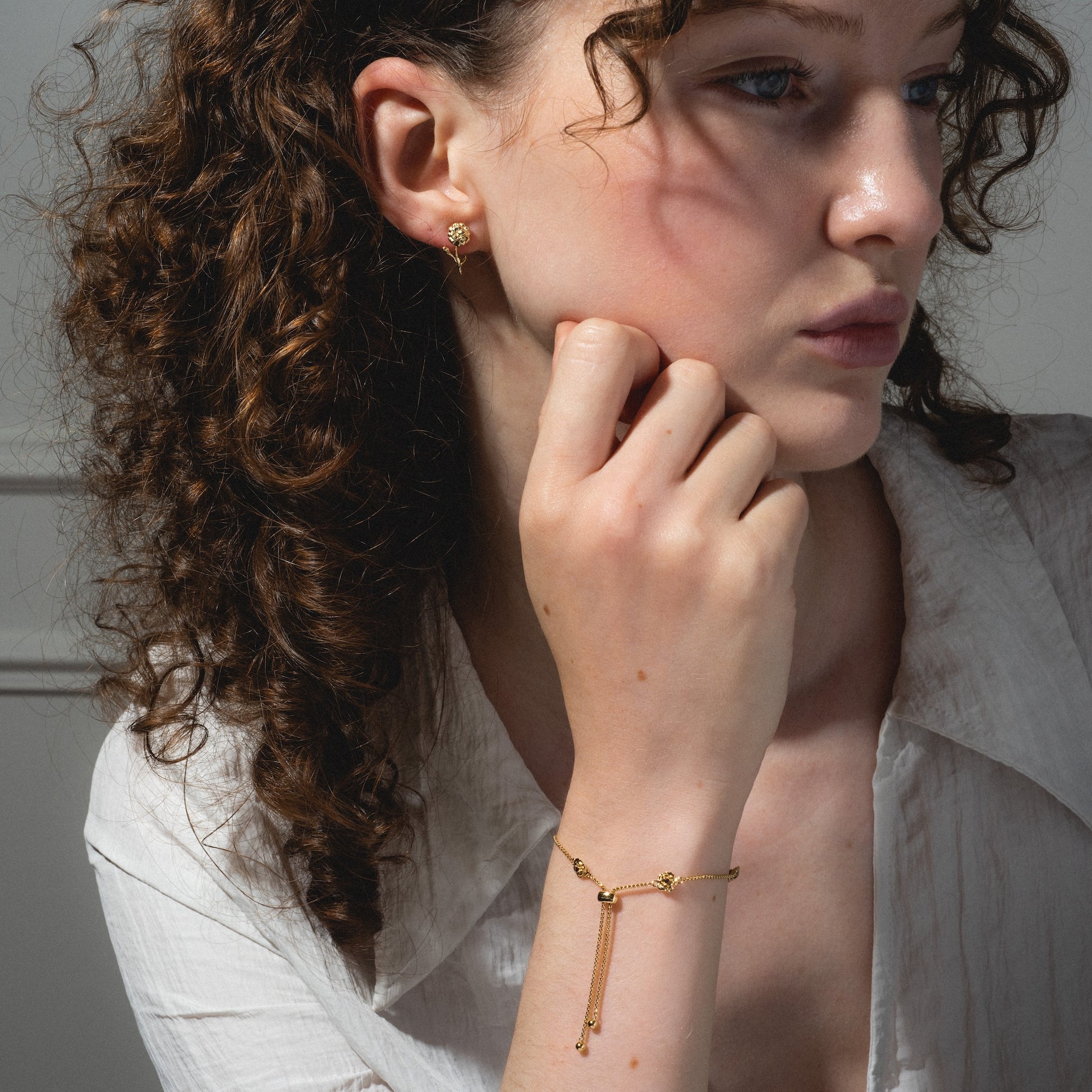 A woman with curly brown hair wears the Birth Flower Gift Set—gold bracelet and floral earrings—resting her chin on her hand and gazing thoughtfully to the side against a neutral background.