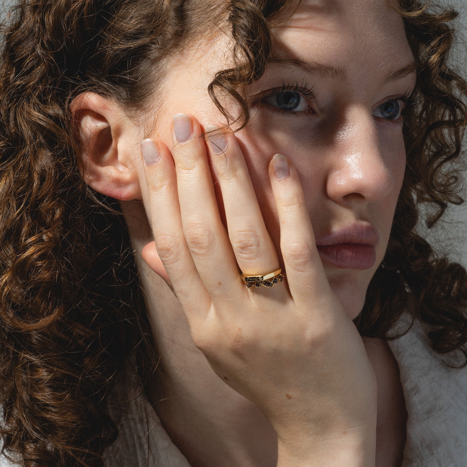A woman with curly brown hair rests her face on her hand, wearing the Tiny Fuck Birthstone Stacking Ring Set in 14k gold plate. The rings complement her natural nails as she gazes thoughtfully into the distance, lit by natural light.