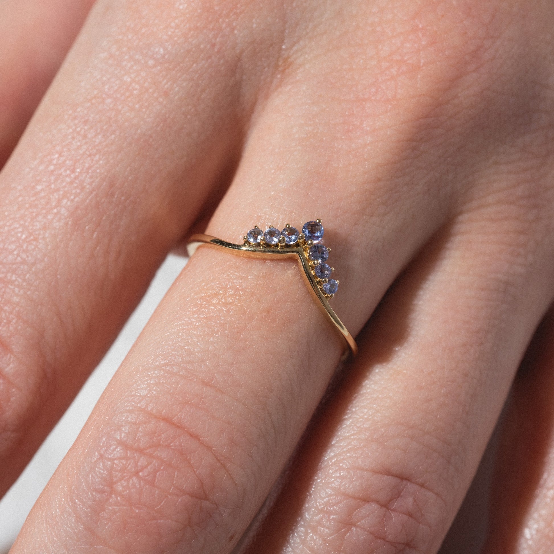 A close-up of a hand wearing the Solid Gold 2025 December Capsule Delicate Arch Tanzanite Stacking Ring, featuring a gold chevron band with small round tanzanite stones, softly lit on the middle finger.