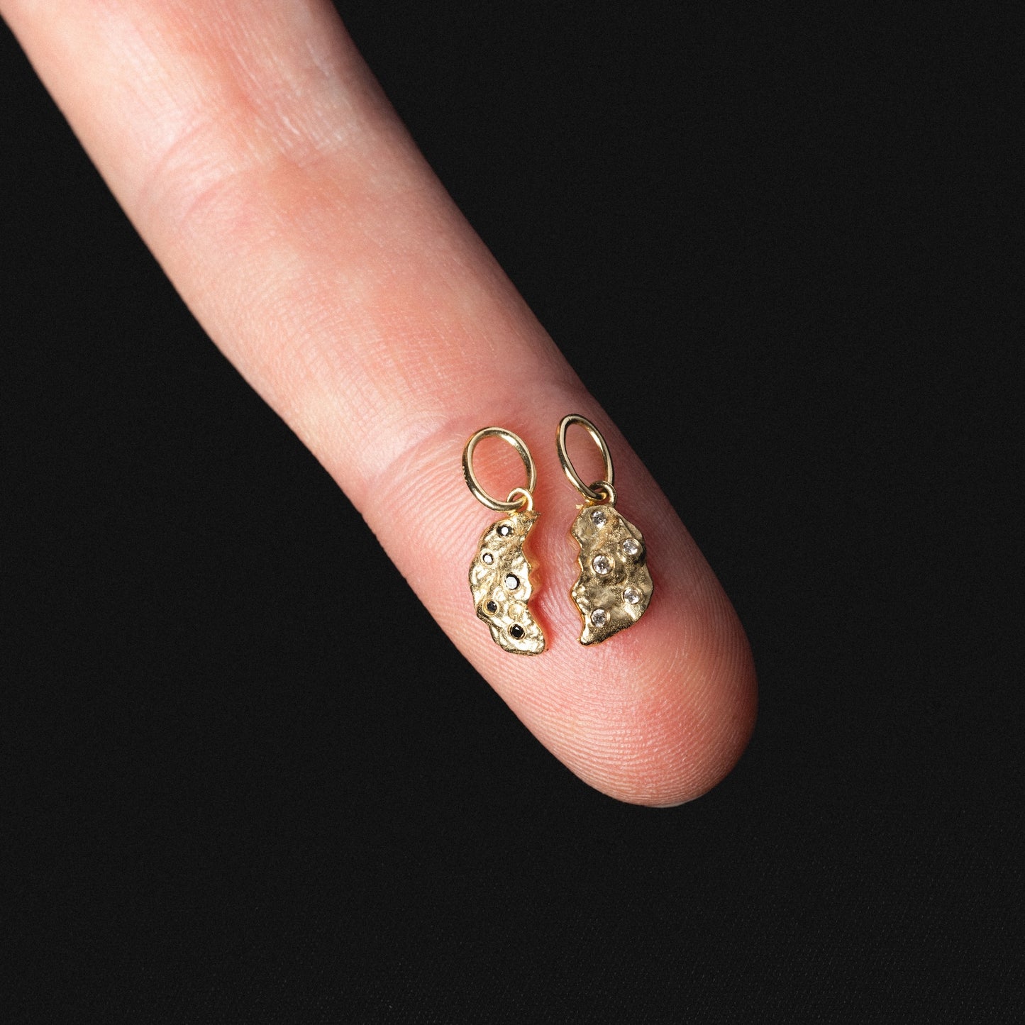 A close-up of a finger holding two Levain Bakery x Local Eclectic Solid Gold Share a Cookie Friendship Charms with textured, irregular shapes and small hoops, displayed against a black background.