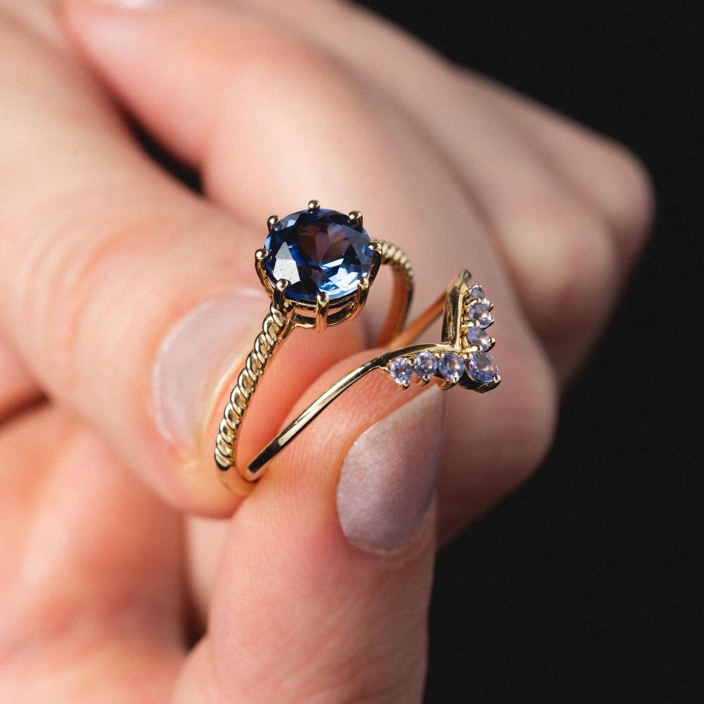 A hand holds two gold rings: one is the Solid Gold 2025 December Capsule Classic Tanzanite Statement Ring with a large blue gemstone in a prong setting; the other features a curved gold band with small clear stones.