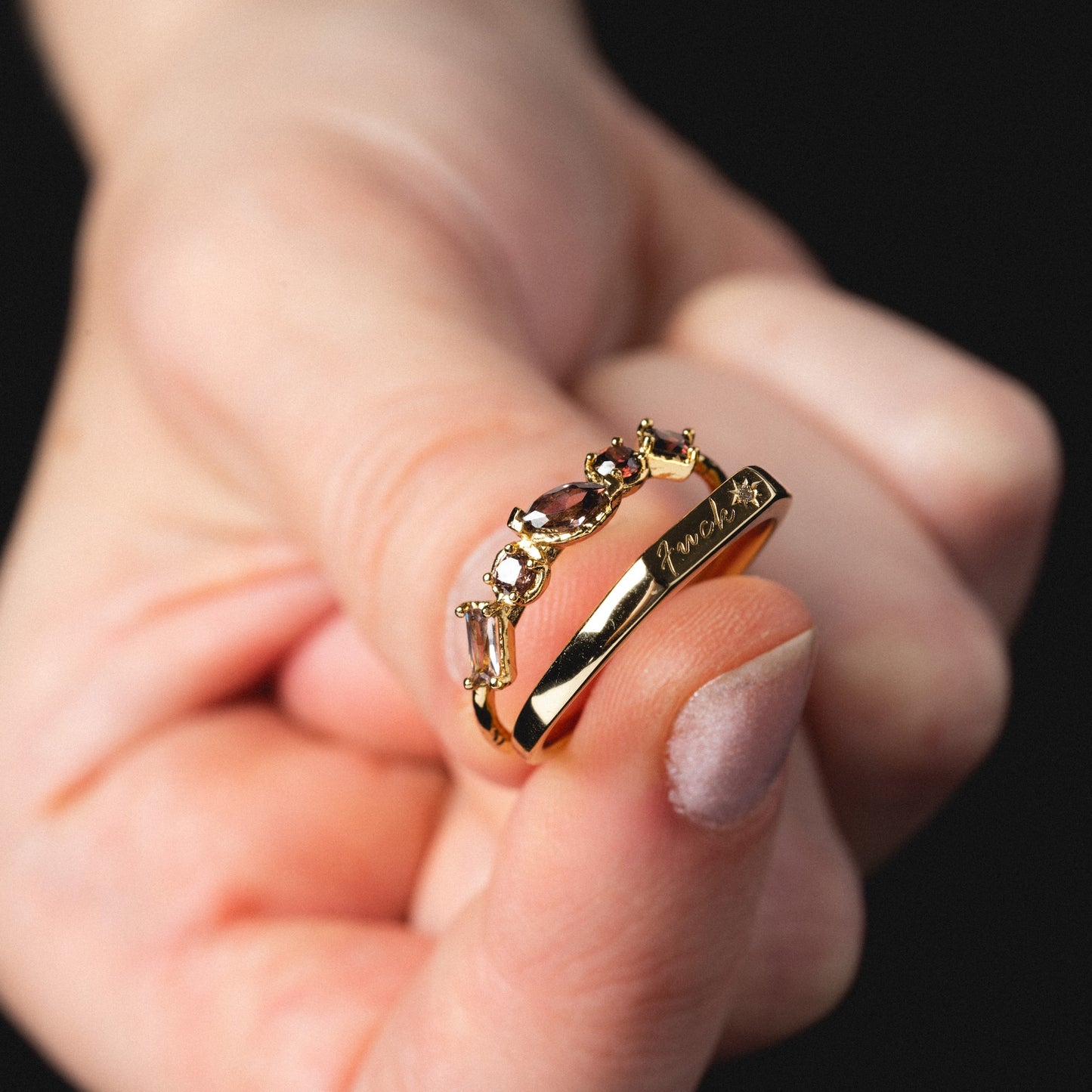 A hand with light nail polish holds the Tiny Fuck Birthstone Stacking Ring Set in 14k yellow gold plating, engraved with “princess,” against a dark background.