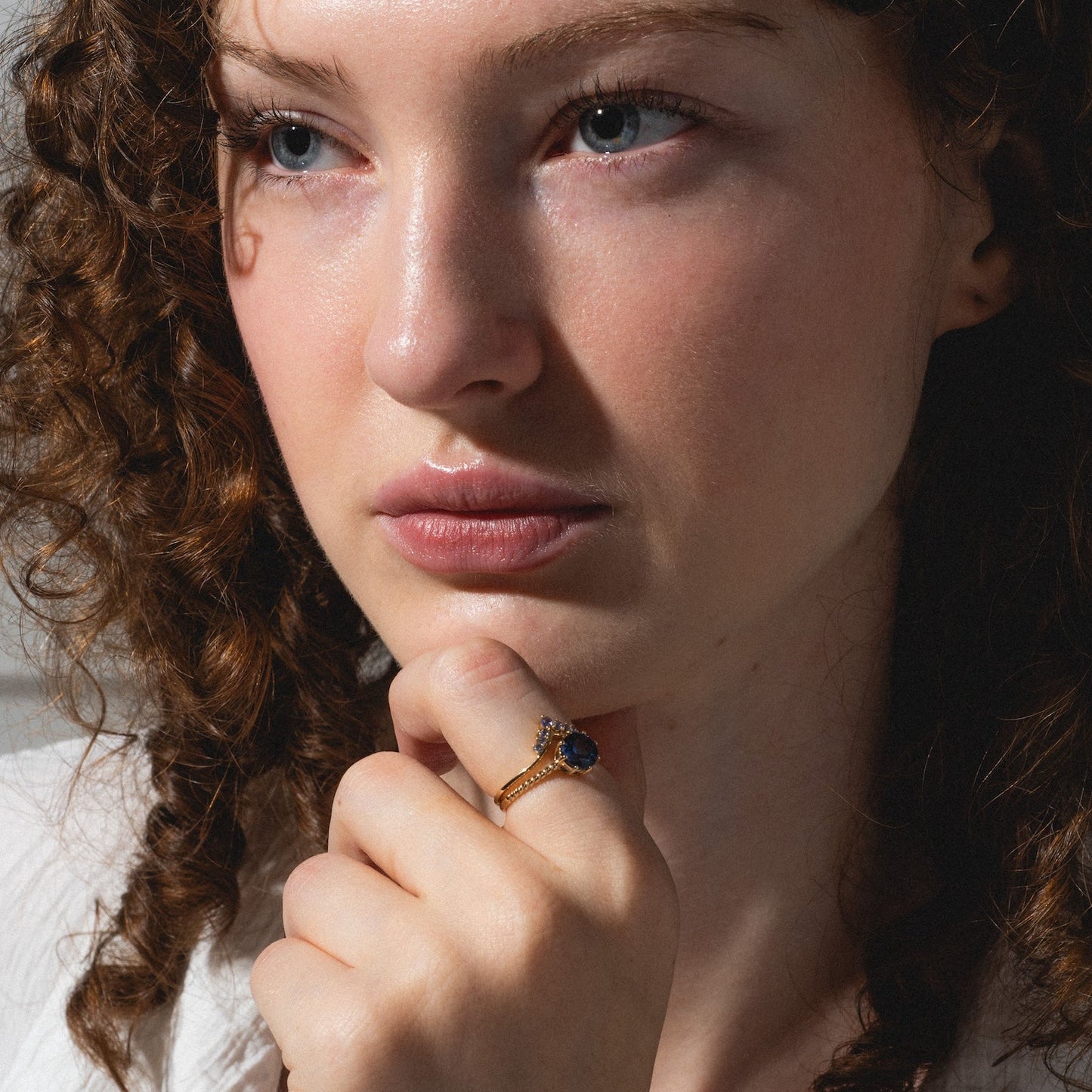 A woman with curly brown hair and fair skin, wearing a white shirt, gazes thoughtfully to the side. Her hand, resting against her chin, displays the Solid Gold 2025 December Capsule Tanzanite Ring Set with Free Gift.