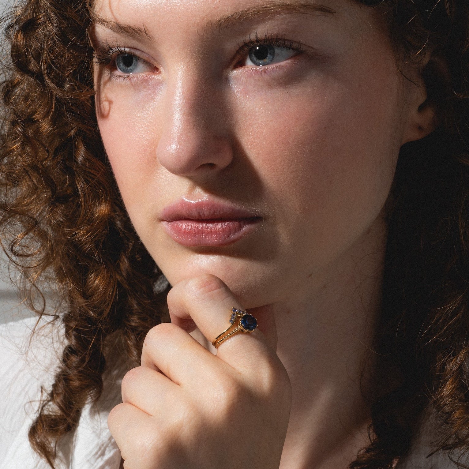 A woman with curly brown hair and fair skin, wearing a white shirt, gazes thoughtfully to the side. Her hand, resting against her chin, displays the Solid Gold 2025 December Capsule Tanzanite Ring Set with Free Gift.