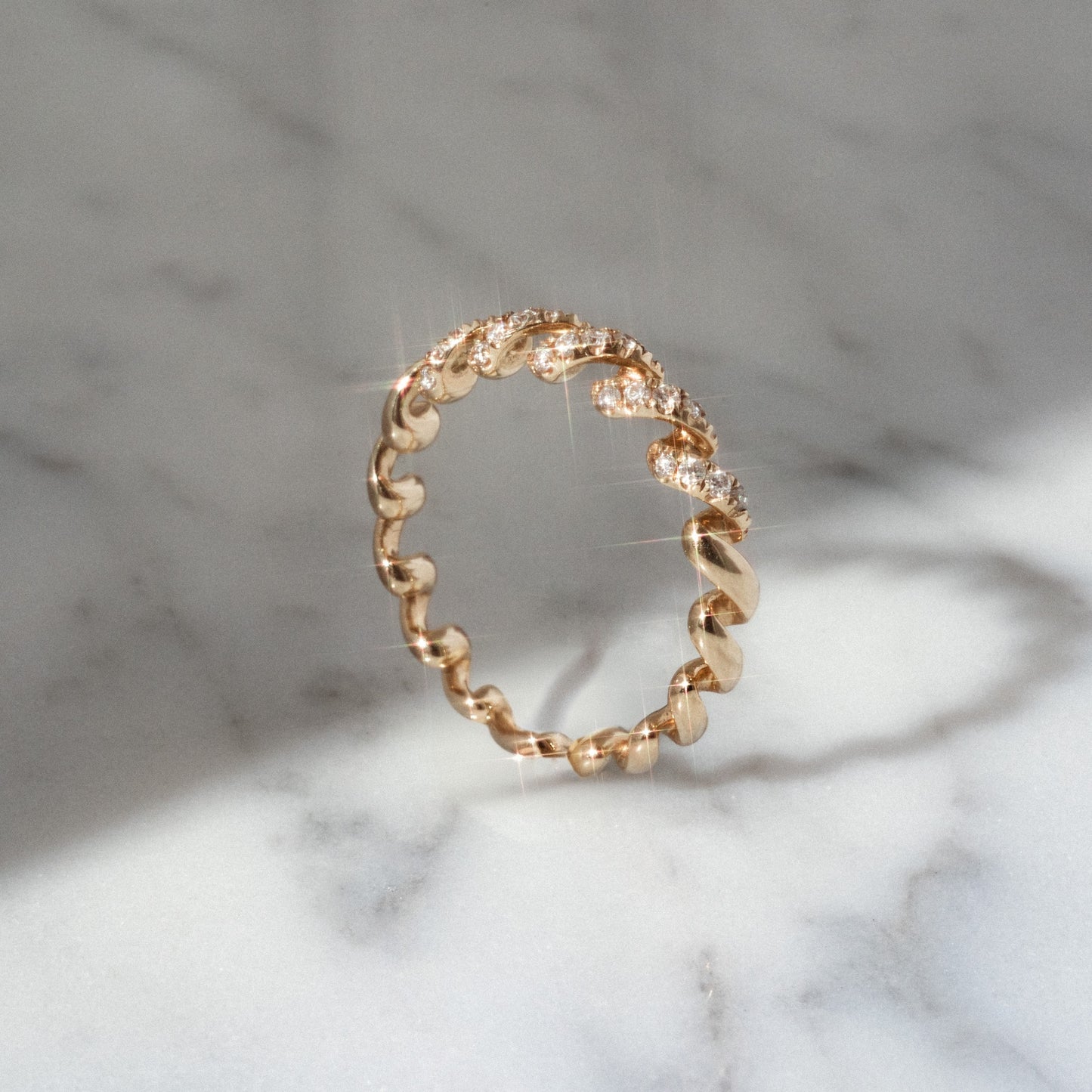 The 14k Fated Twist Diamond Ring features a twisted, wavy gold design with sparkling diamonds on top, beautifully displayed on a white marble surface under soft natural light.