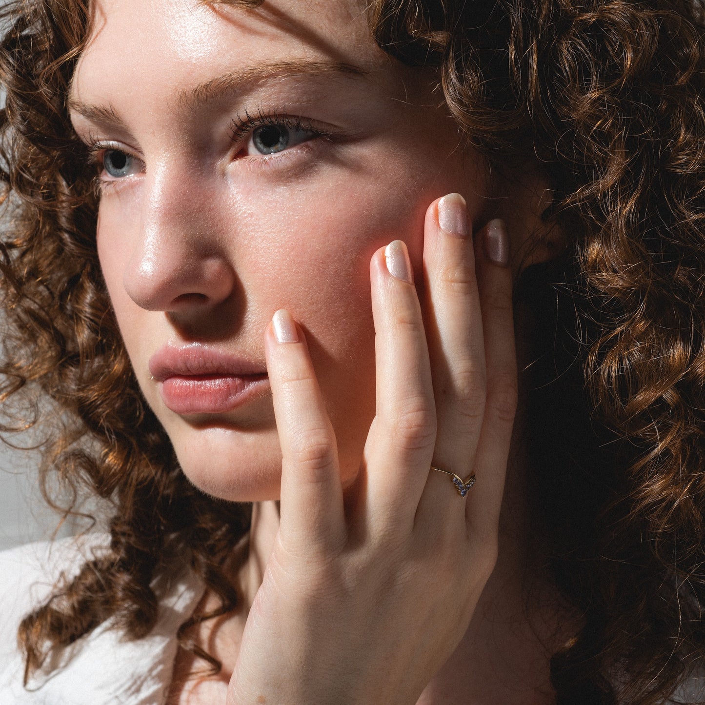 A person with curly brown hair gazes thoughtfully, gently resting their hand—adorned with the Solid Gold 2025 December Capsule Delicate Arch Tanzanite Stacking Ring—on their cheek. Soft natural light enhances their clear skin and subtle makeup.
