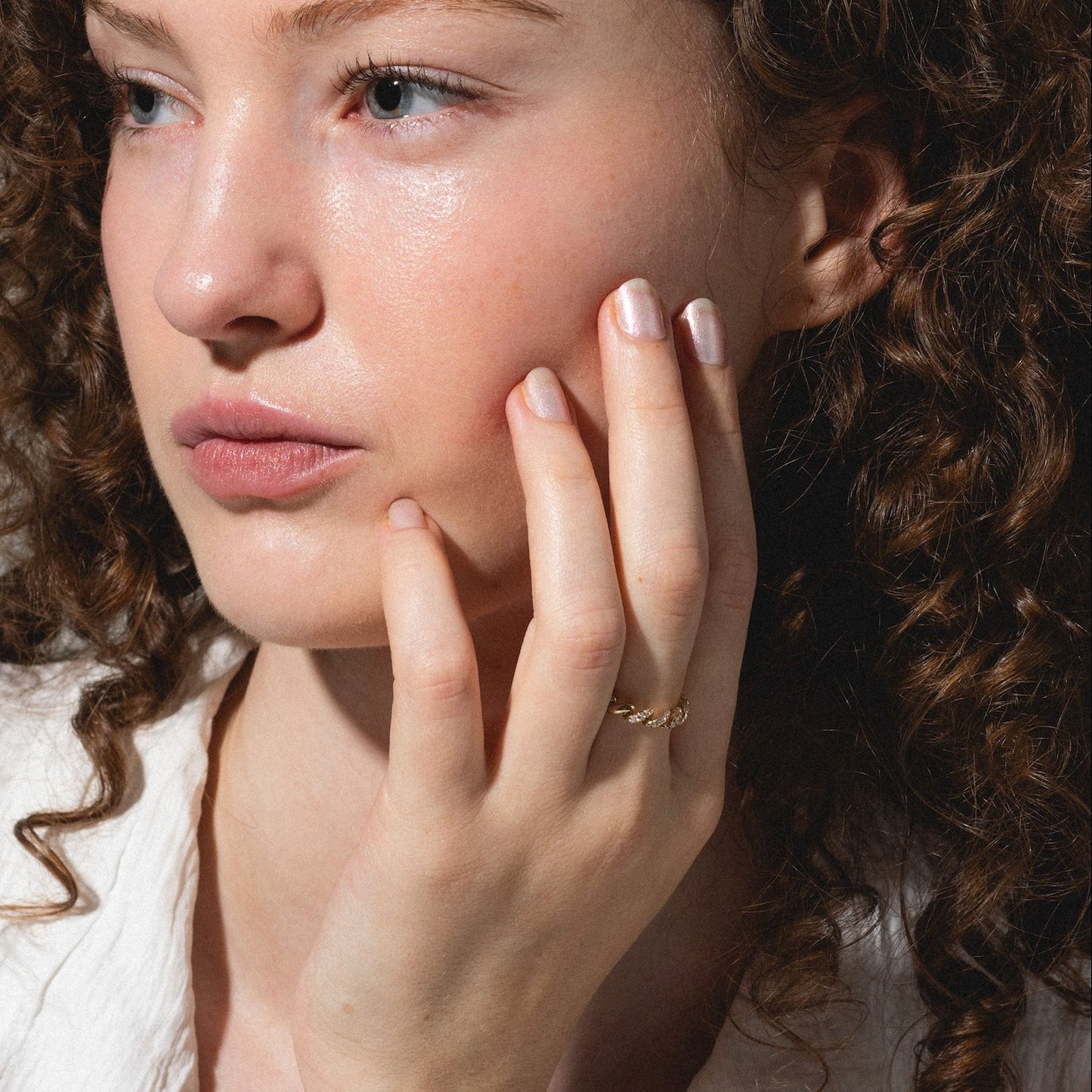 A woman with curly brown hair and light skin gazes thoughtfully to the side, resting her hand on her cheek. She wears a white shirt and the 14k Fated Twist Diamond Ring, her expression calm and contemplative.
