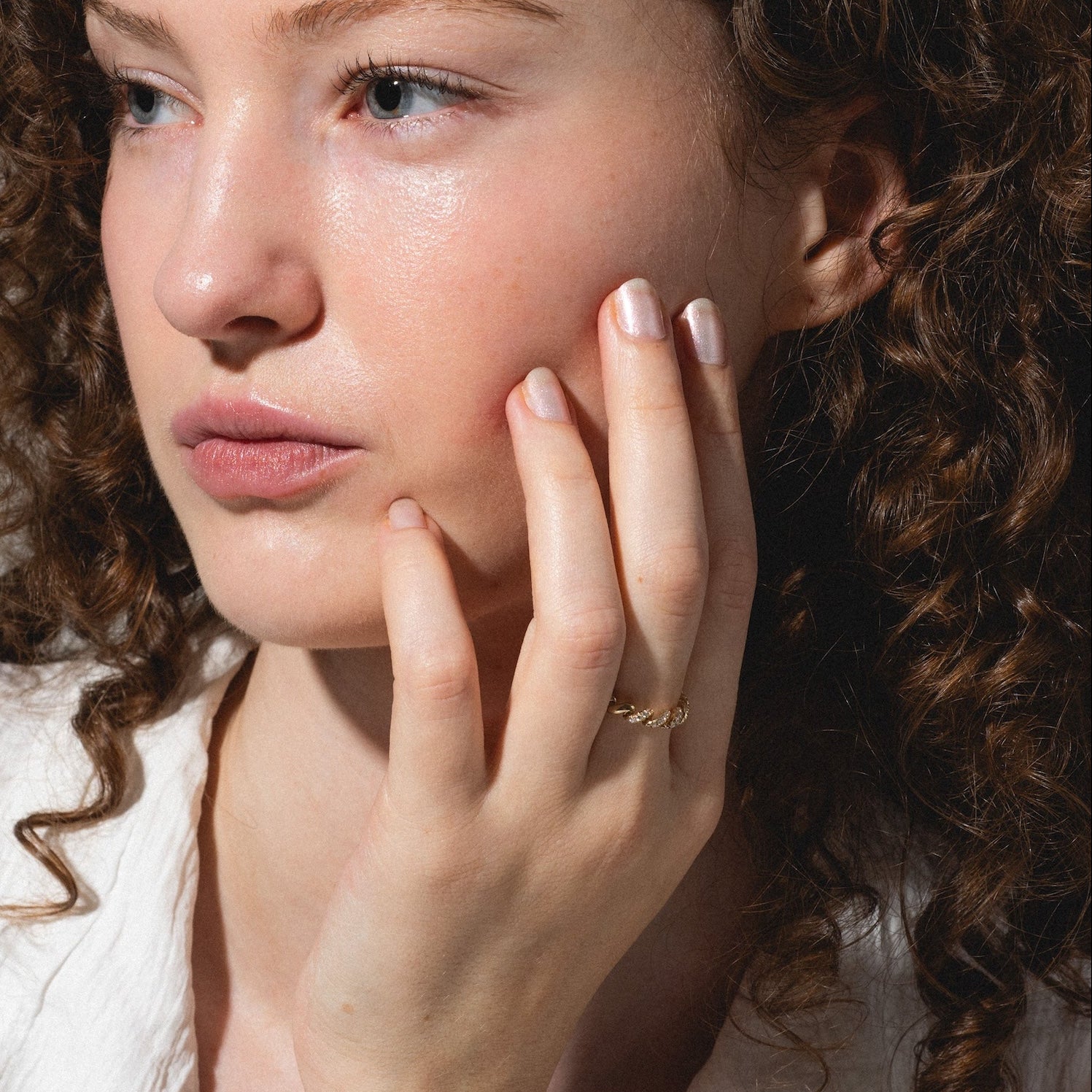 A woman with curly brown hair and light skin gazes thoughtfully to the side, resting her hand on her cheek. She wears a white shirt and the 14k Fated Twist Diamond Ring, her expression calm and contemplative.