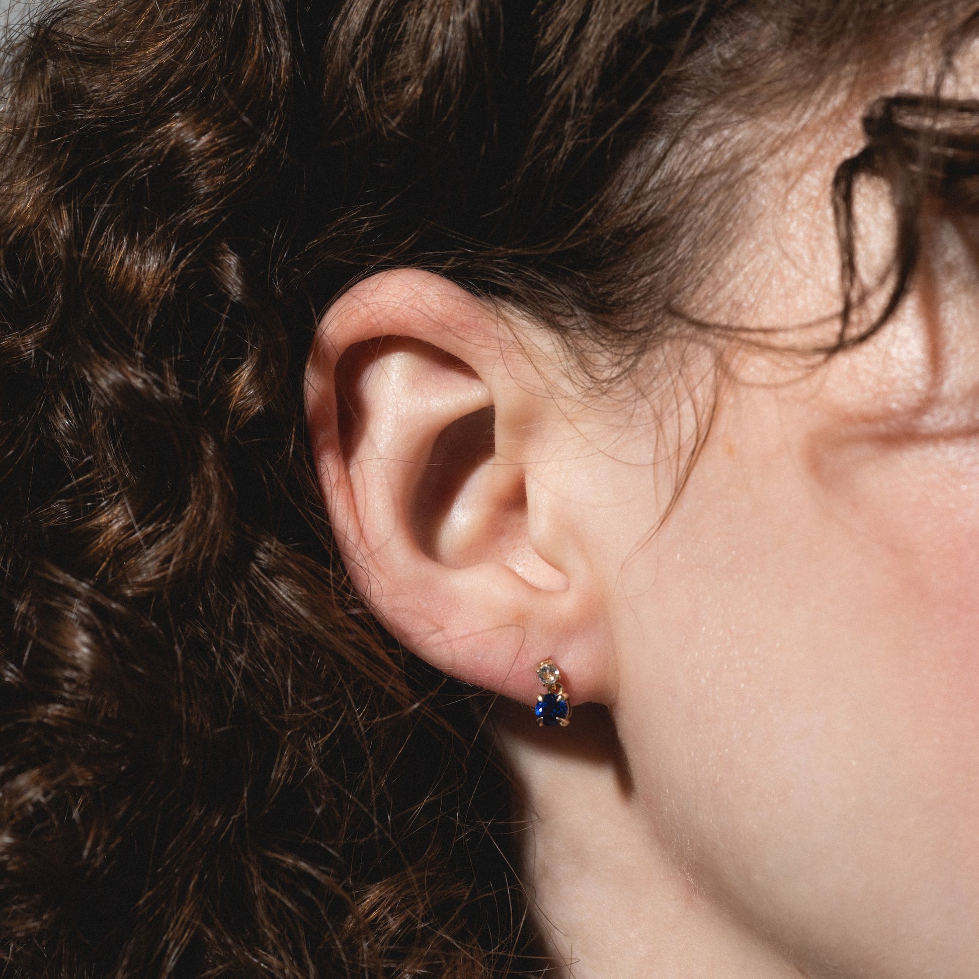Close-up of a woman's ear with curly brown hair, wearing Solid Gold 2025 December Capsule Classic Tanzanite Stud Earrings with a blue gemstone and clear stone. Lighting highlights the texture of her hair and skin.