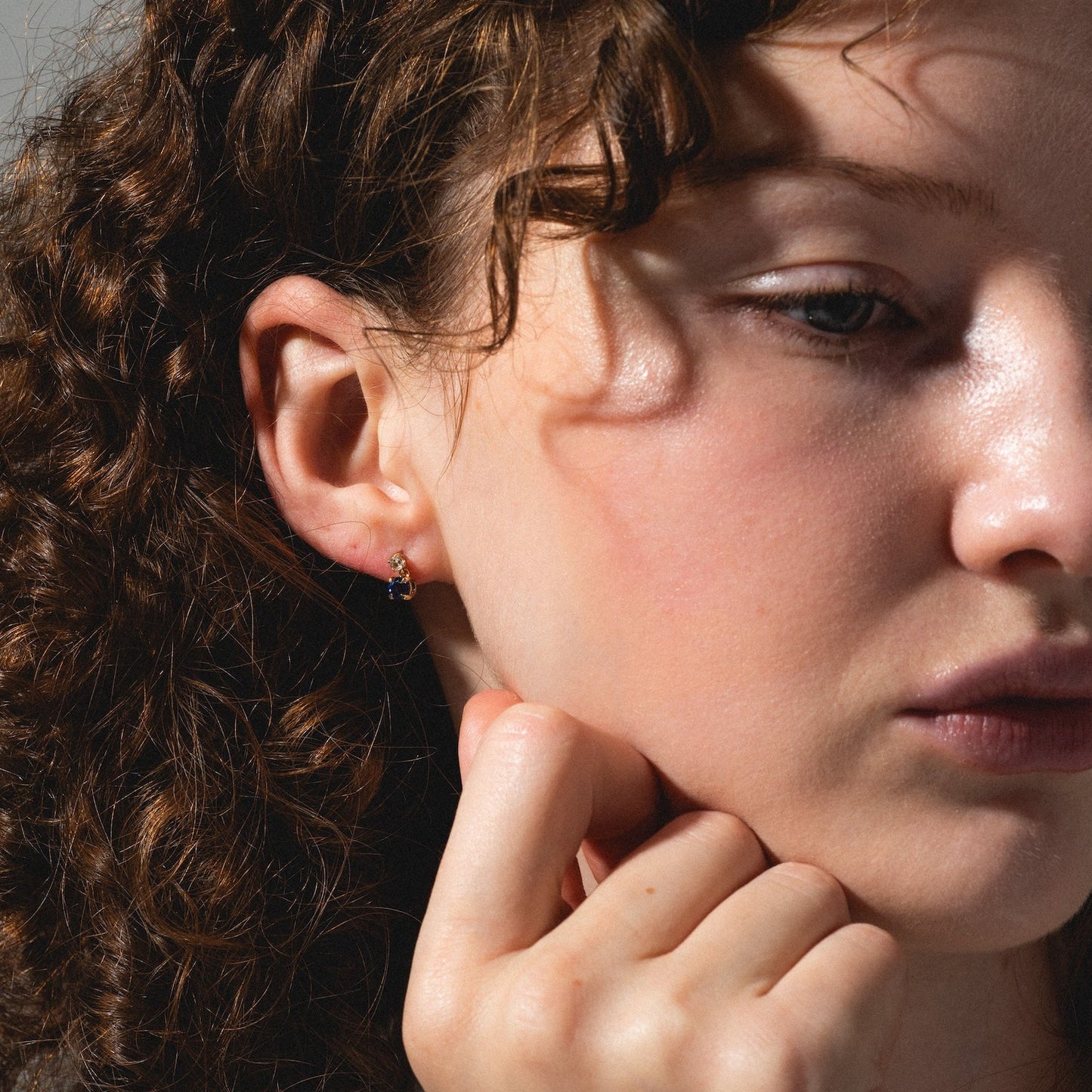 A woman with curly brown hair gazes downward thoughtfully, resting her chin on her hand. She wears Solid Gold 2025 December Capsule Classic Tanzanite Stud Earrings and has a calm, pensive expression in soft natural light.