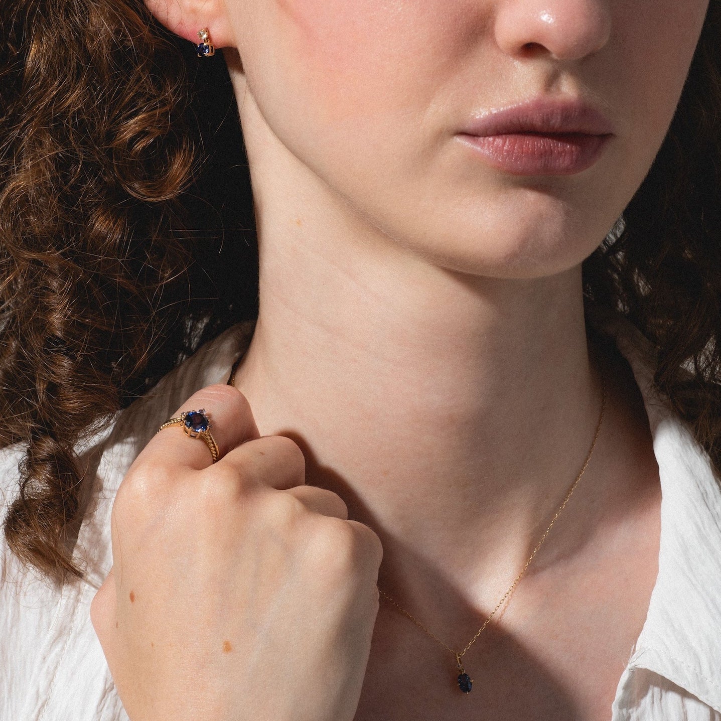 A woman with curly brown hair wears a white textured shirt and the Solid Gold 2025 December Capsule Classic Tanzanite Pendant Necklace. She also has a matching gold ring, small gold earring, and looks ahead with a neutral expression.