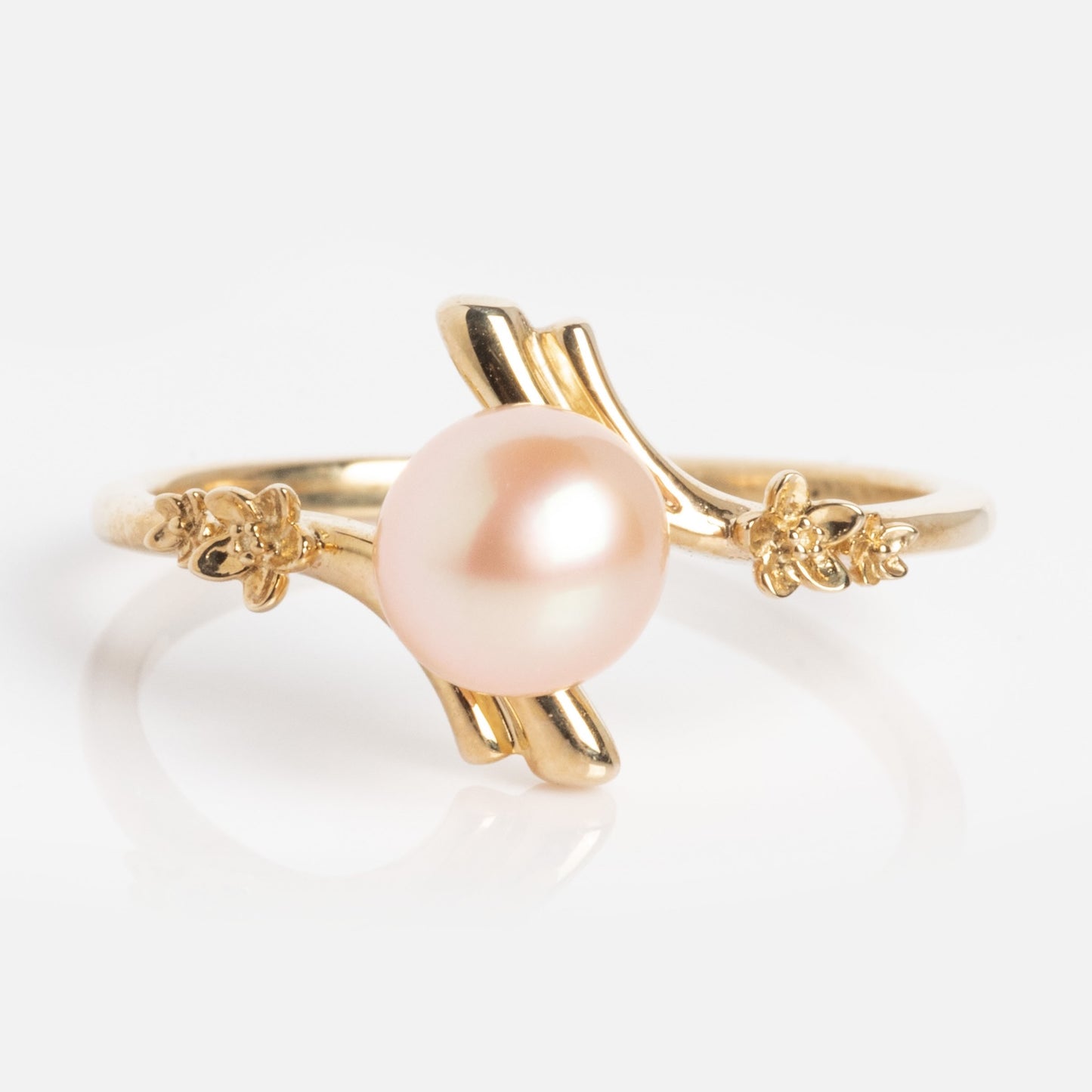 The 10k Forget Me Not Pink Pearl Bypass Ring showcases a round, light pink pearl at the center, accented by delicate gold floral details on each side, all set against a white background.