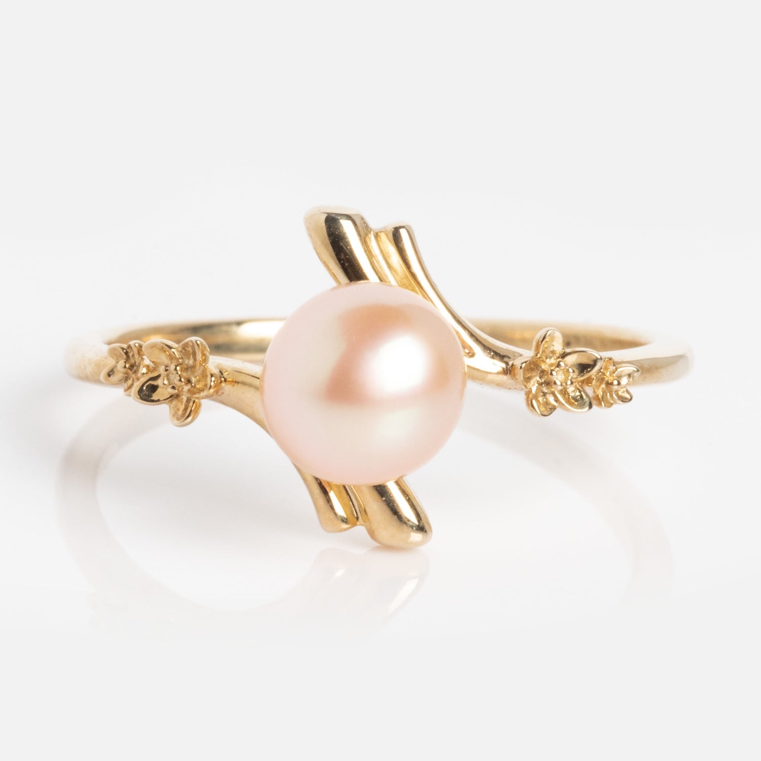 The 10k Forget Me Not Pink Pearl Bypass Ring showcases a round, light pink pearl at the center, accented by delicate gold floral details on each side, all set against a white background.