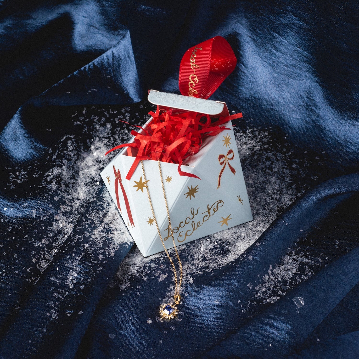 The Limited Edition 2025 Holiday Ornament Gift Packaging features a decorative box with red shredded paper, gold stars, and red ribbon on blue satin, topped with a gold necklace and artificial snow for a festive touch.
