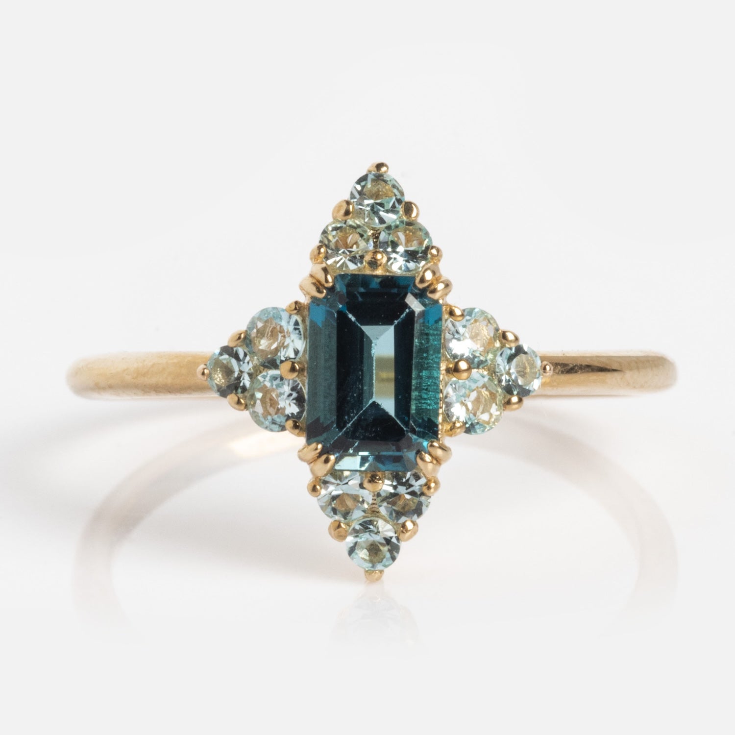 The 10k Snow Queen Blue Topaz Cluster Ring features a rectangular dark blue topaz at the center, encircled by symmetrically arranged clusters of small light blue stones.
