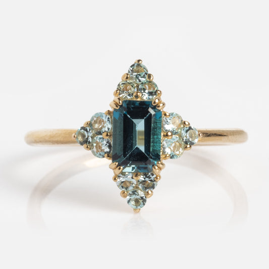 The 10k Snow Queen Blue Topaz Cluster Ring features a rectangular dark blue topaz at the center, encircled by symmetrically arranged clusters of small light blue stones.