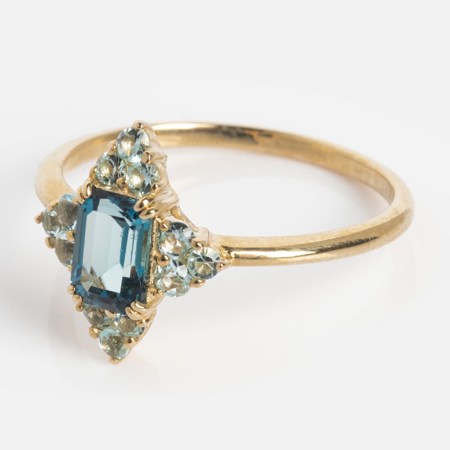 The 10k Snow Queen Blue Topaz Cluster Ring features a rectangular blue topaz center stone, surrounded by smaller light blue gems on a plain gold band, showcased against a white background.
