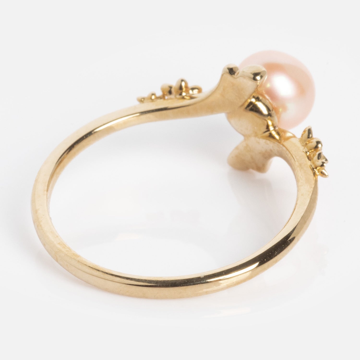 The 10k Forget Me Not Pink Pearl Bypass Ring features a pink pearl atop a delicate, nature-inspired band with leaf-like accents, shown on a white background.