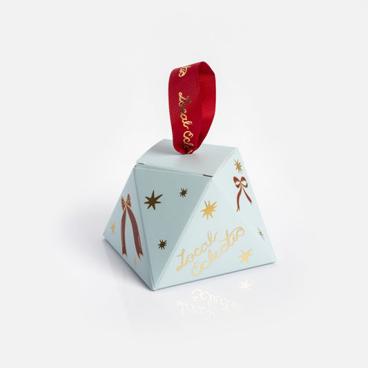 The Limited Edition 2025 Holiday Ornament Gift Packaging is a light blue, pyramid-shaped box with gold stars and bows, a red ribbon loop at the top, and “Local Eclectic” in gold text.