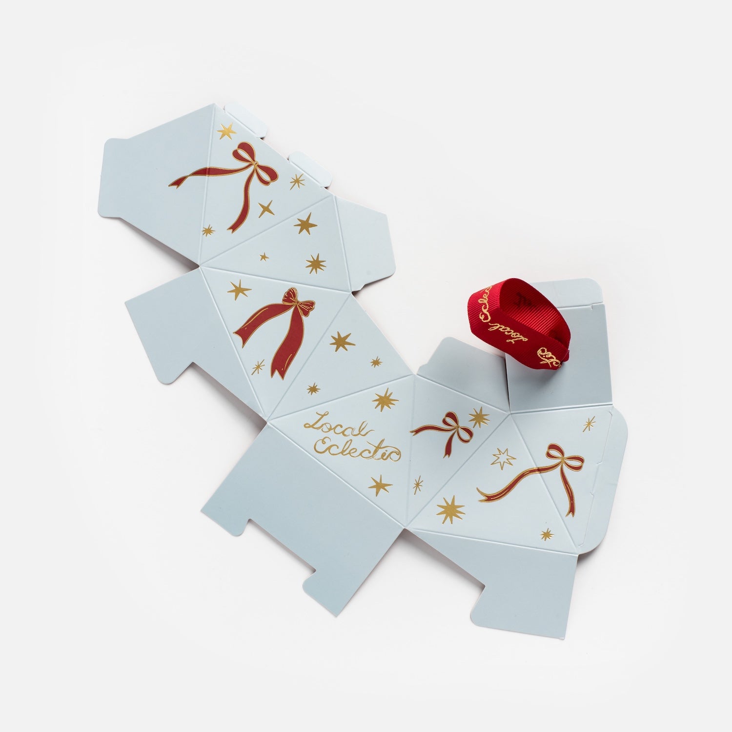 The Limited Edition 2025 Holiday Ornament Gift Packaging features a flat, unfolded light gray box with gold stars, red bows, "local eclectic" text, and a looped red ribbon with gold lettering placed on top.