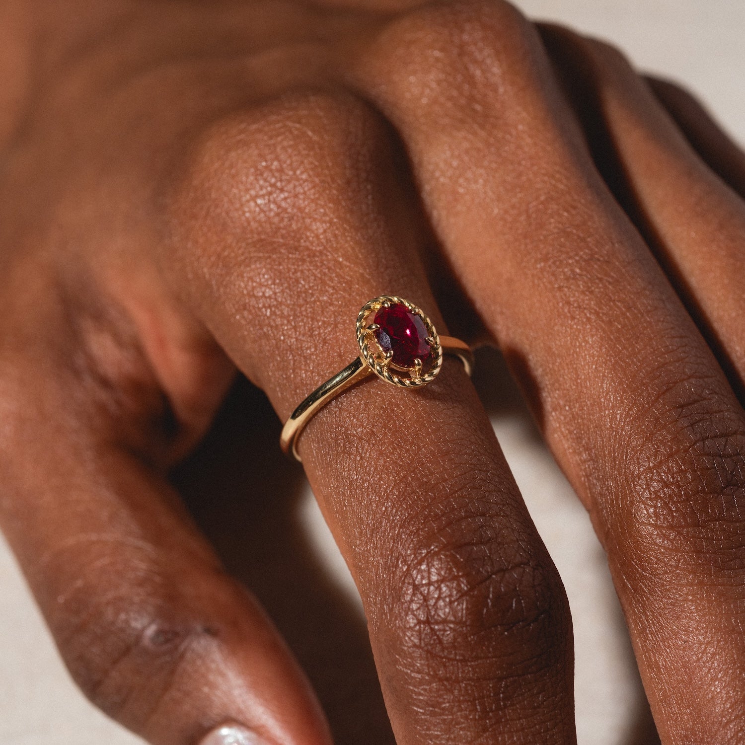 A hand displays the 10k Lucky In Red Ruby Halo Ring featuring an oval red gemstone in a decorative bezel, set against a neutral background.