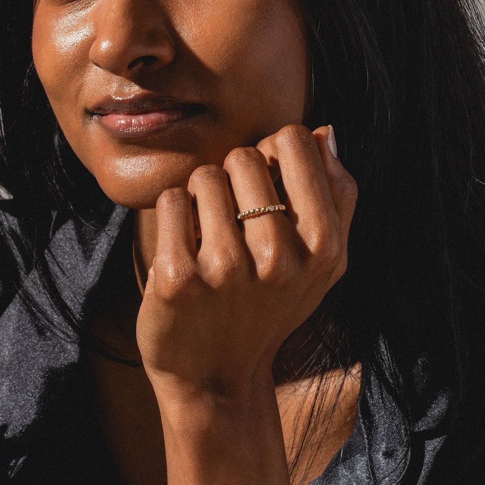 A woman with long dark hair wearing a black satin shirt rests her chin on her hand, displaying the 10k Hidden Message Morse Code Diamond Band. She gazes at the camera with a gentle smile in soft, natural light.